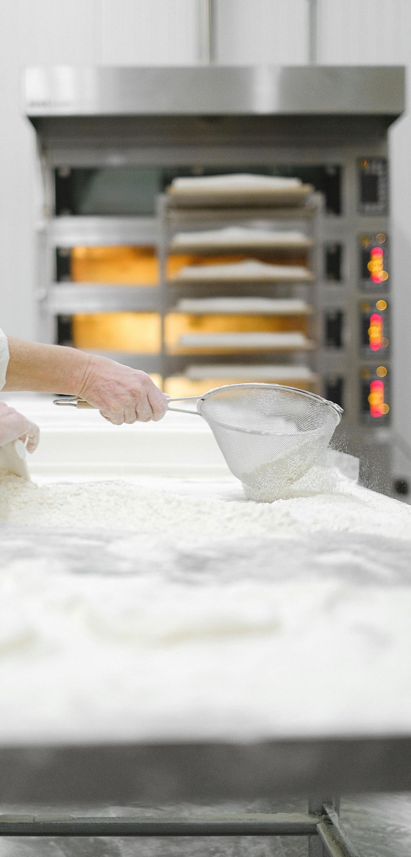 A person uses a sieve to dust flour onto a white table in a kitchen with a stainless steel industrial oven in the back.