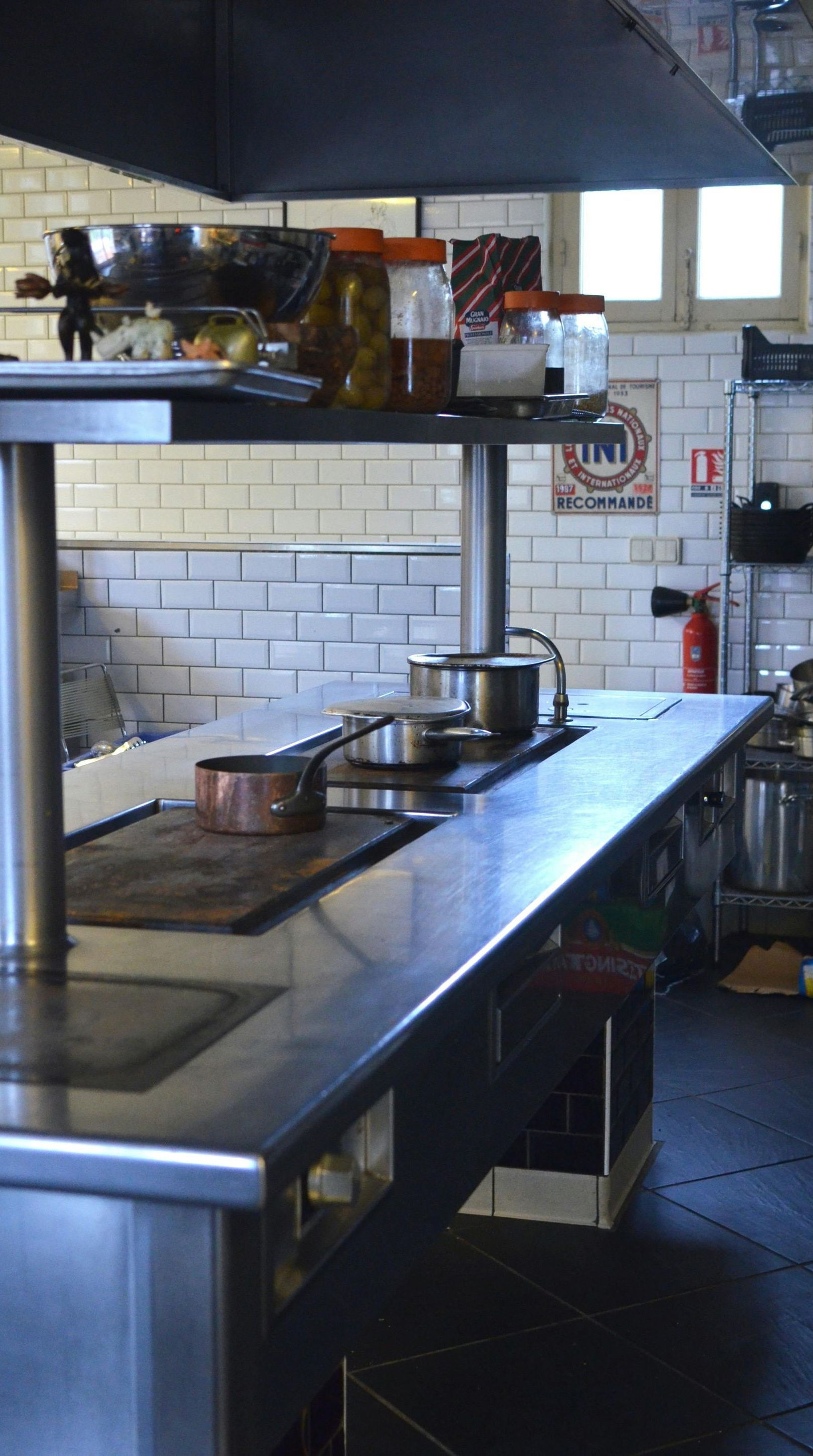 Stainless steel commercial kitchen with stovetop, pots, and jars on shelf; white tile wall.