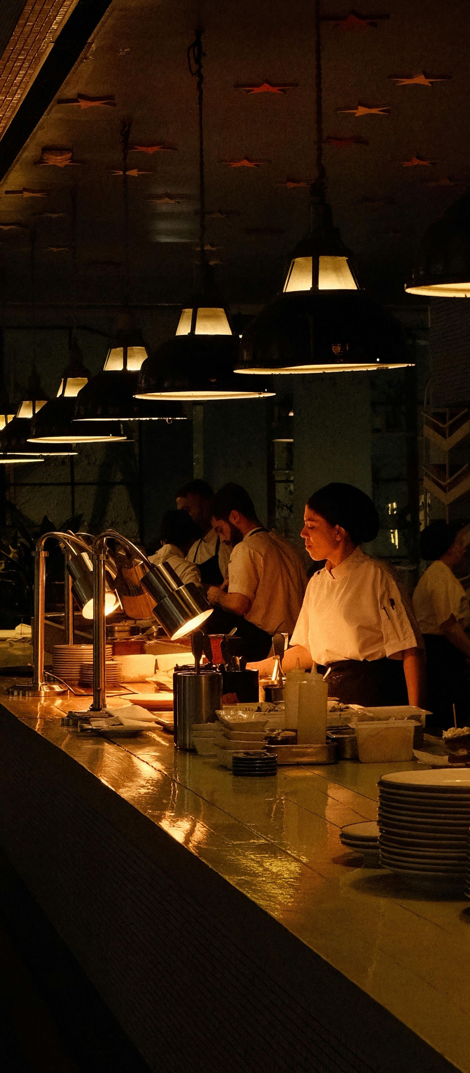 Dimly lit buffet counter with people serving food under hanging pendant lights