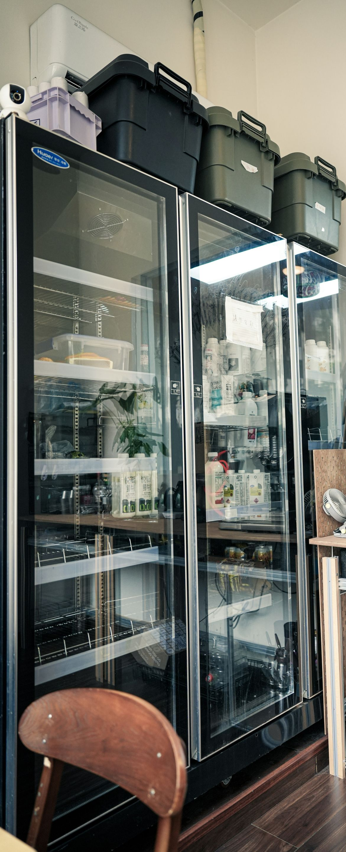 Small café corner with glass display fridges, stacked containers, and a wooden chair by the counter.