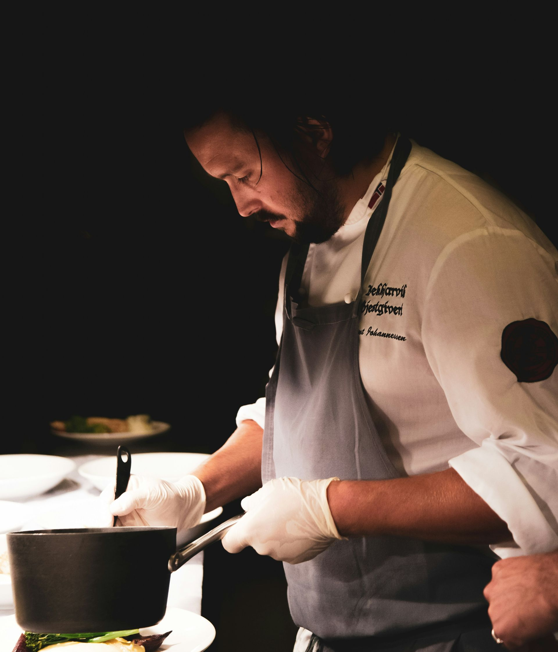 Chef in white jacket and dark apron cooking over a pot on a stovetop in a dim kitchen