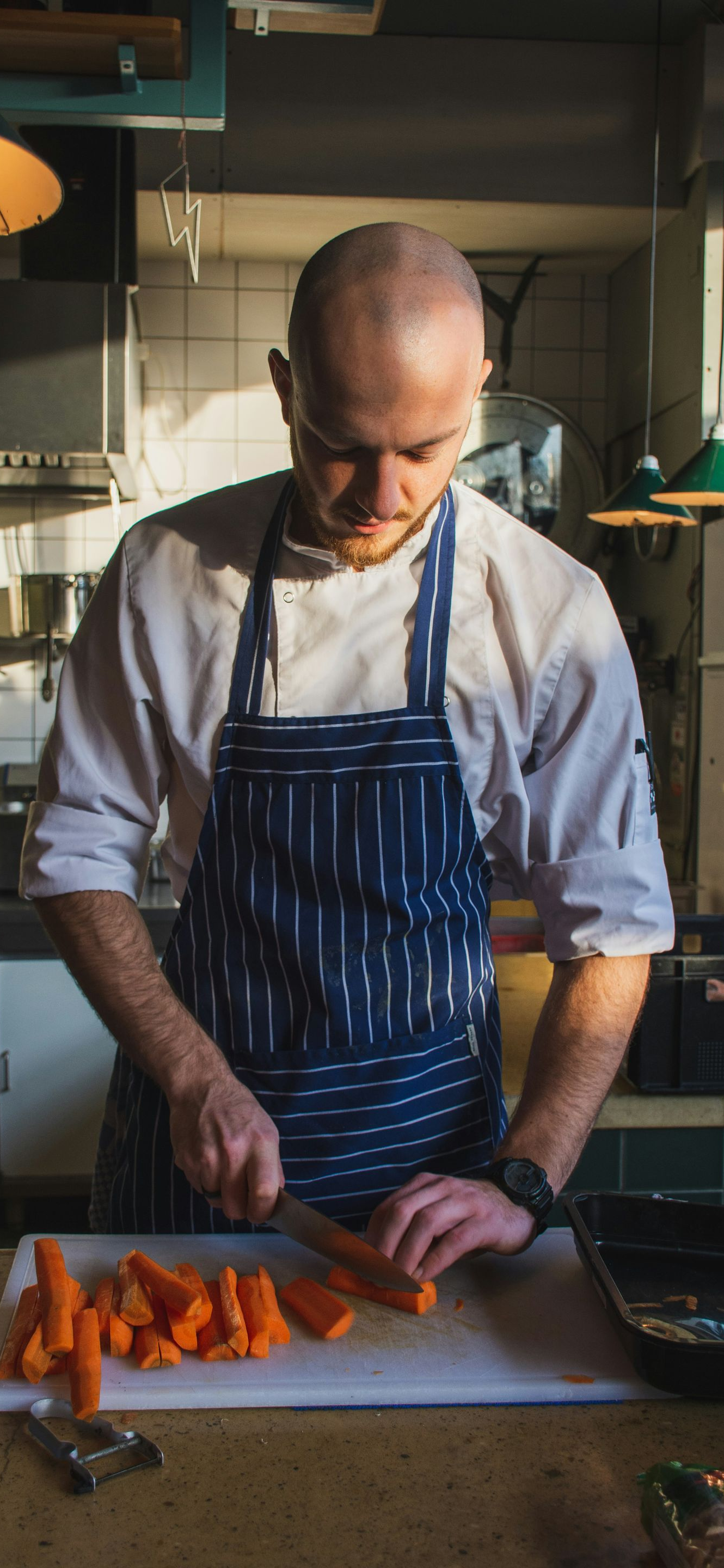 Chef in a striped apron slicing orange vegetables on a cutting board in a kitchen.