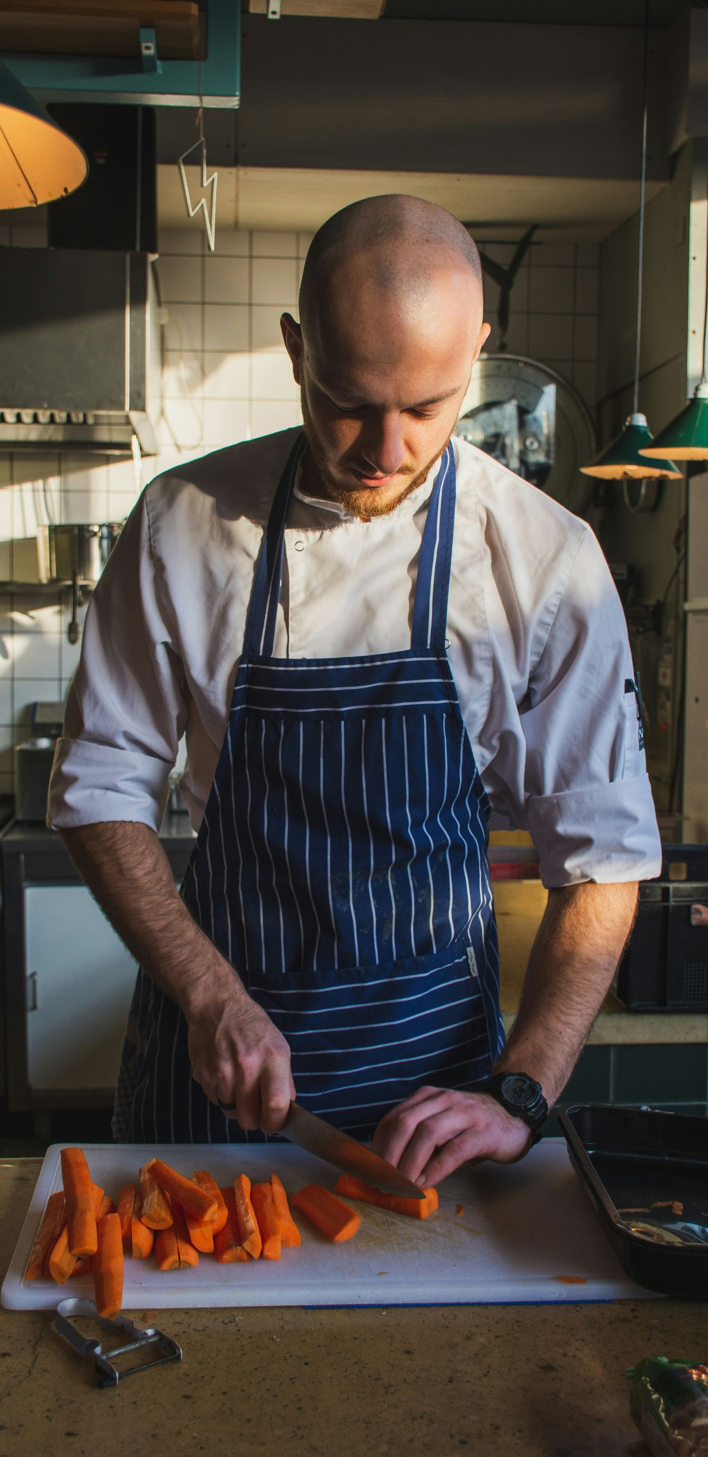 Chef slicing carrots on a cutting board in a sunlit kitchen, wearing a striped apron.