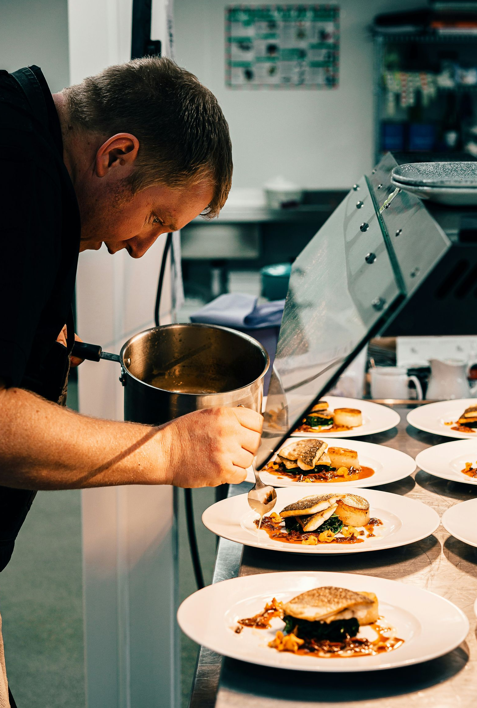 A chef in a professional kitchen drizzles sauce onto several plates of plated fish entrées arranged on a counter.