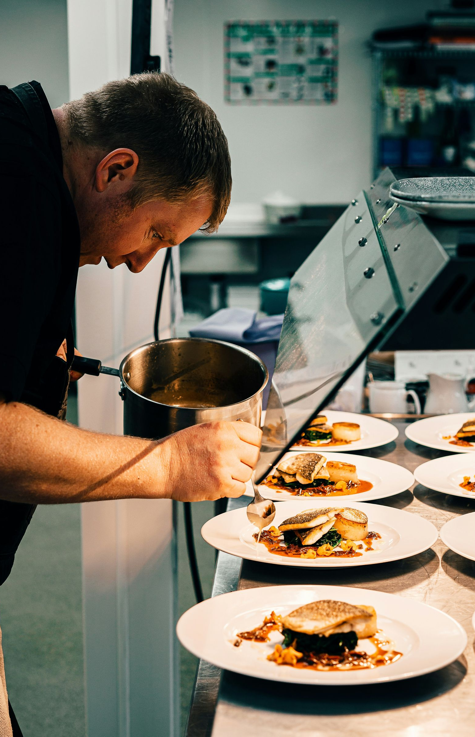A chef in a professional kitchen drizzles sauce onto several plates of plated fish entrées arranged on a counter.