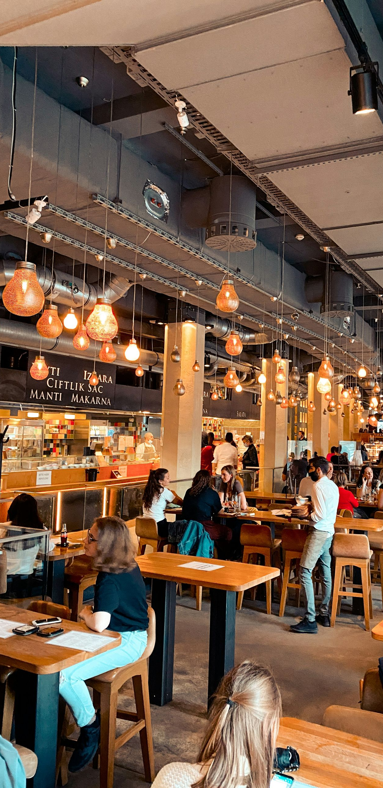 Patrons sit at wooden tables in a busy, industrial-style restaurant with warm overhead lighting.