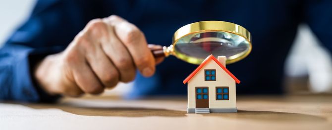 Een hand houdt een vergrootglas boven een klein modelhuis op een tafel, wat duidt op een woninginspectie of een beoordeling van het onroerend goed.