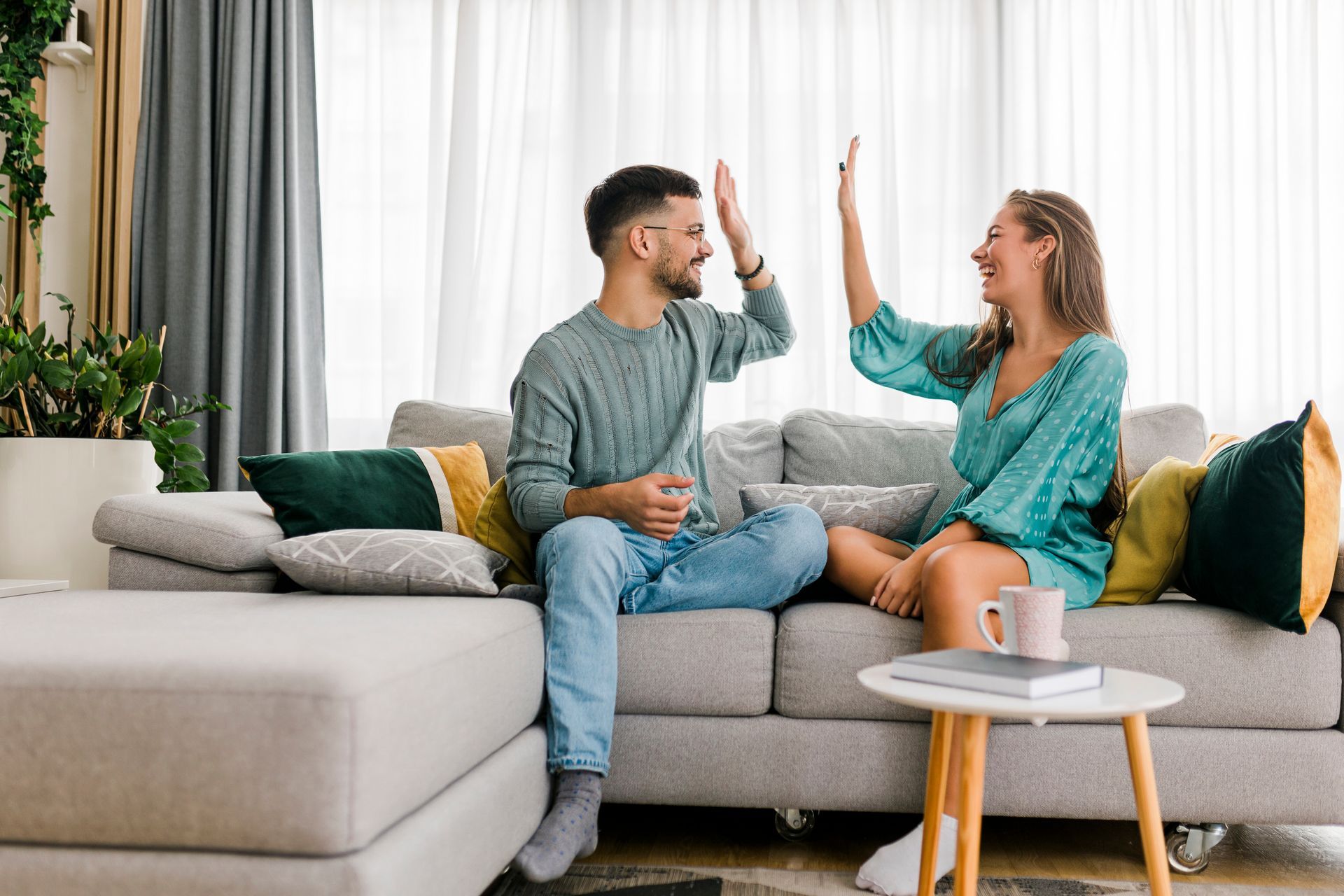 Twee mensen zitten op een bank en geven elkaar een high-five in een lichte woonkamer met kussens en een klein tafeltje.