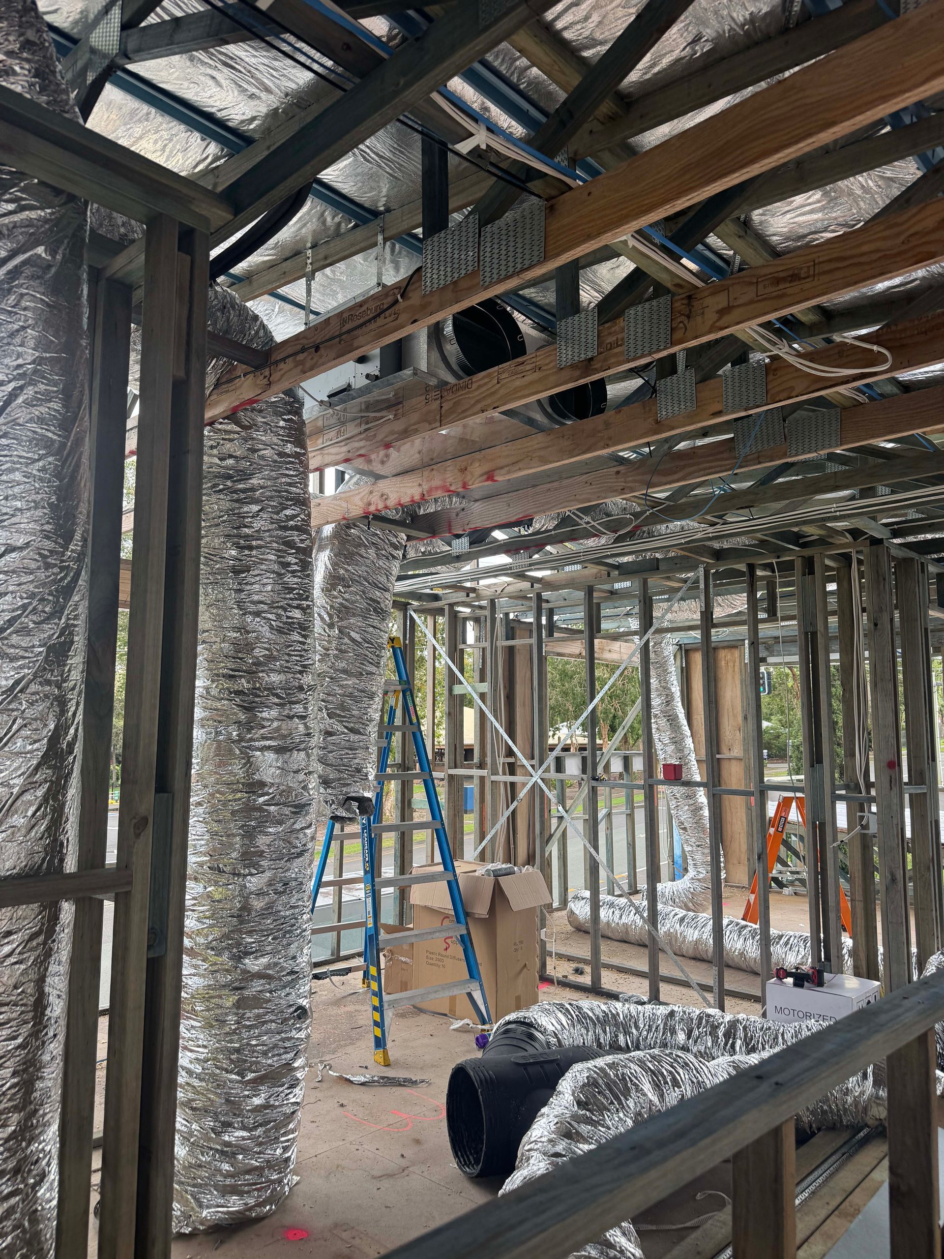 Interior of a building under construction, showing exposed framing, insulation, and HVAC ductwork. A blue ladder is present.