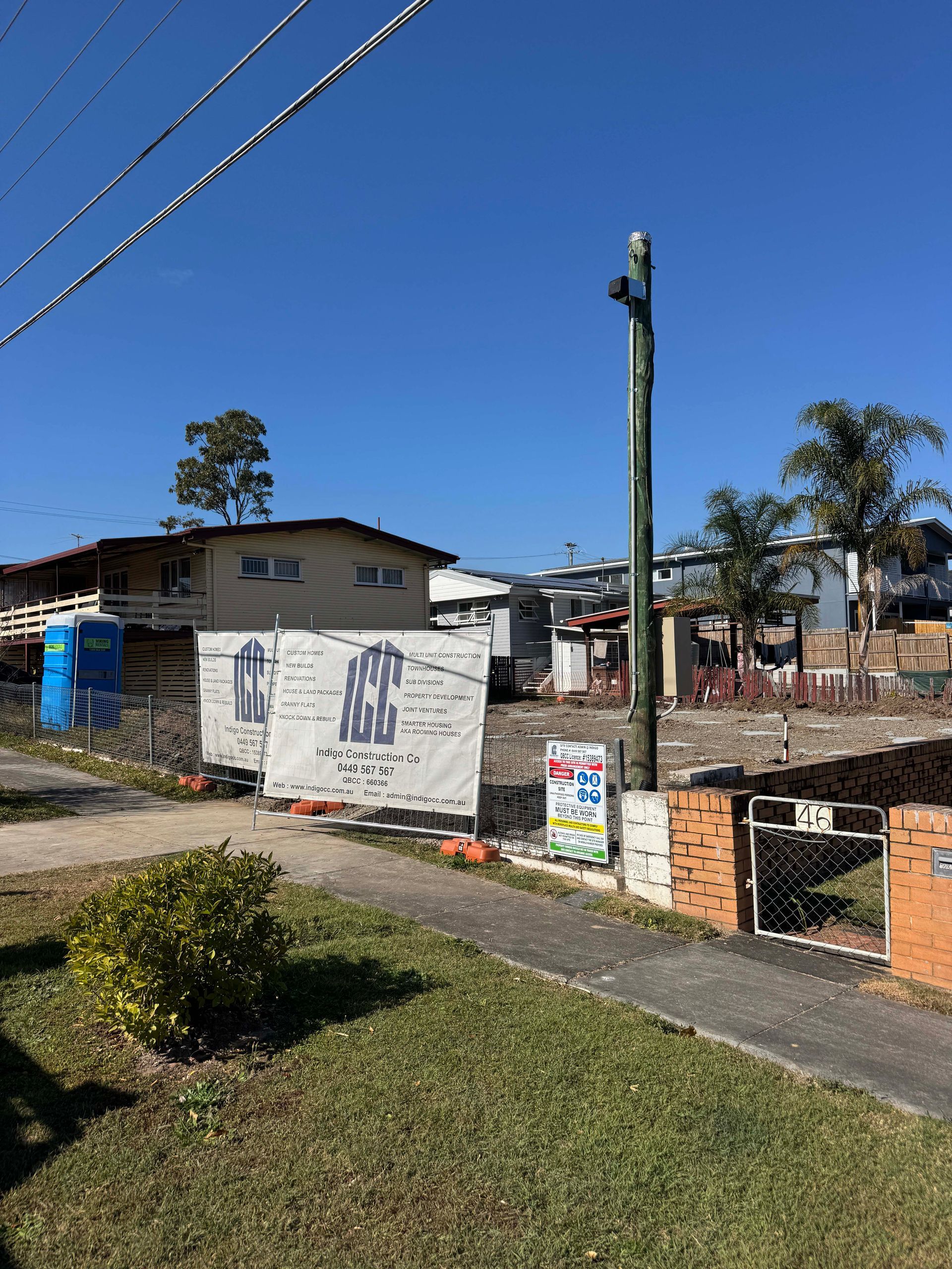Construction site with white banners and a power pole on a sunny day. Houses are visible in the background.