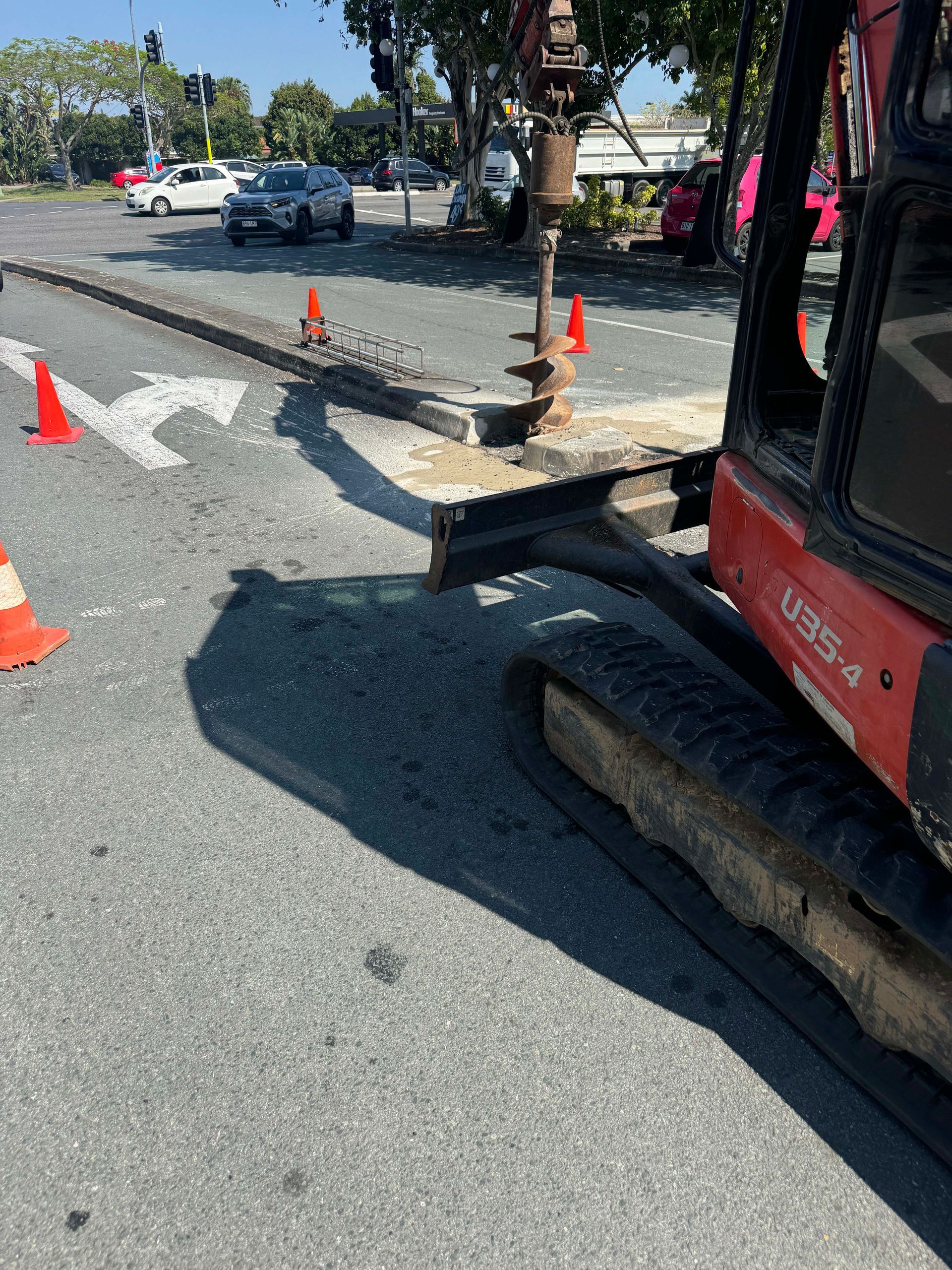 A small red excavator drills into the asphalt road near a crosswalk with orange cones. Cars drive in the background.