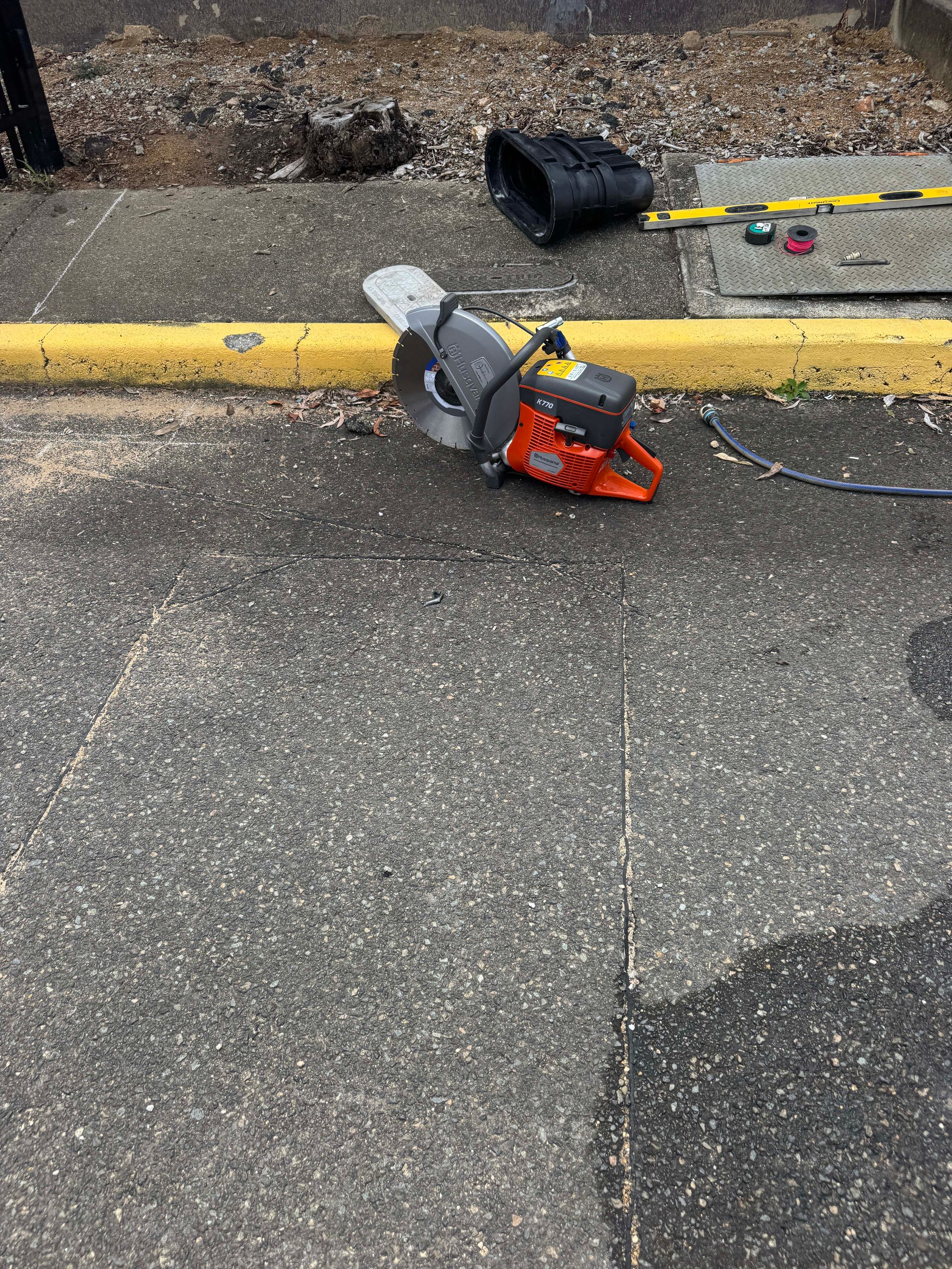 An orange and gray cut-off saw rests on a concrete surface, next to a yellow curb and black pipe.