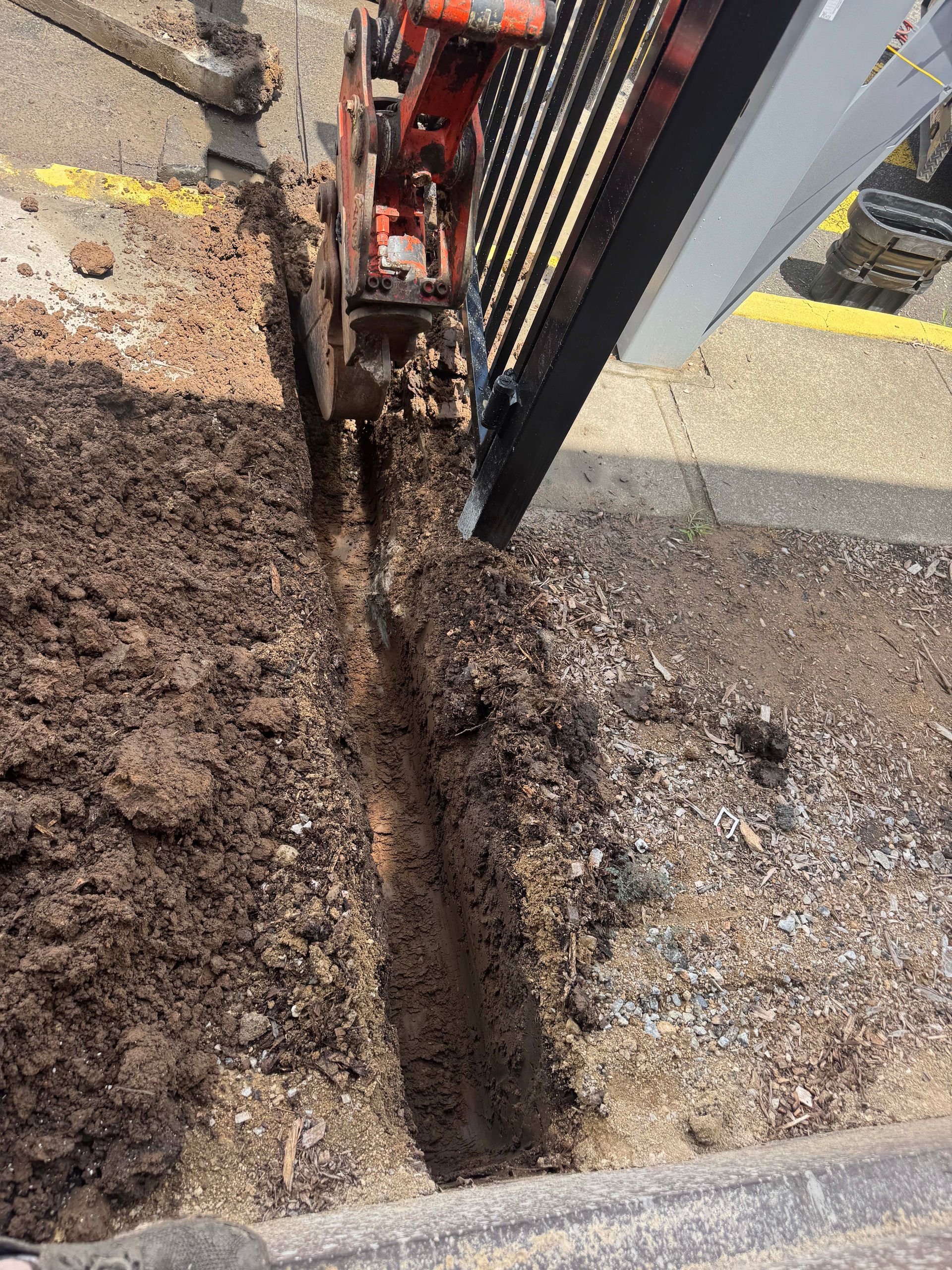 A small excavator digs a trench in the ground next to a paved area, likely for utility work.