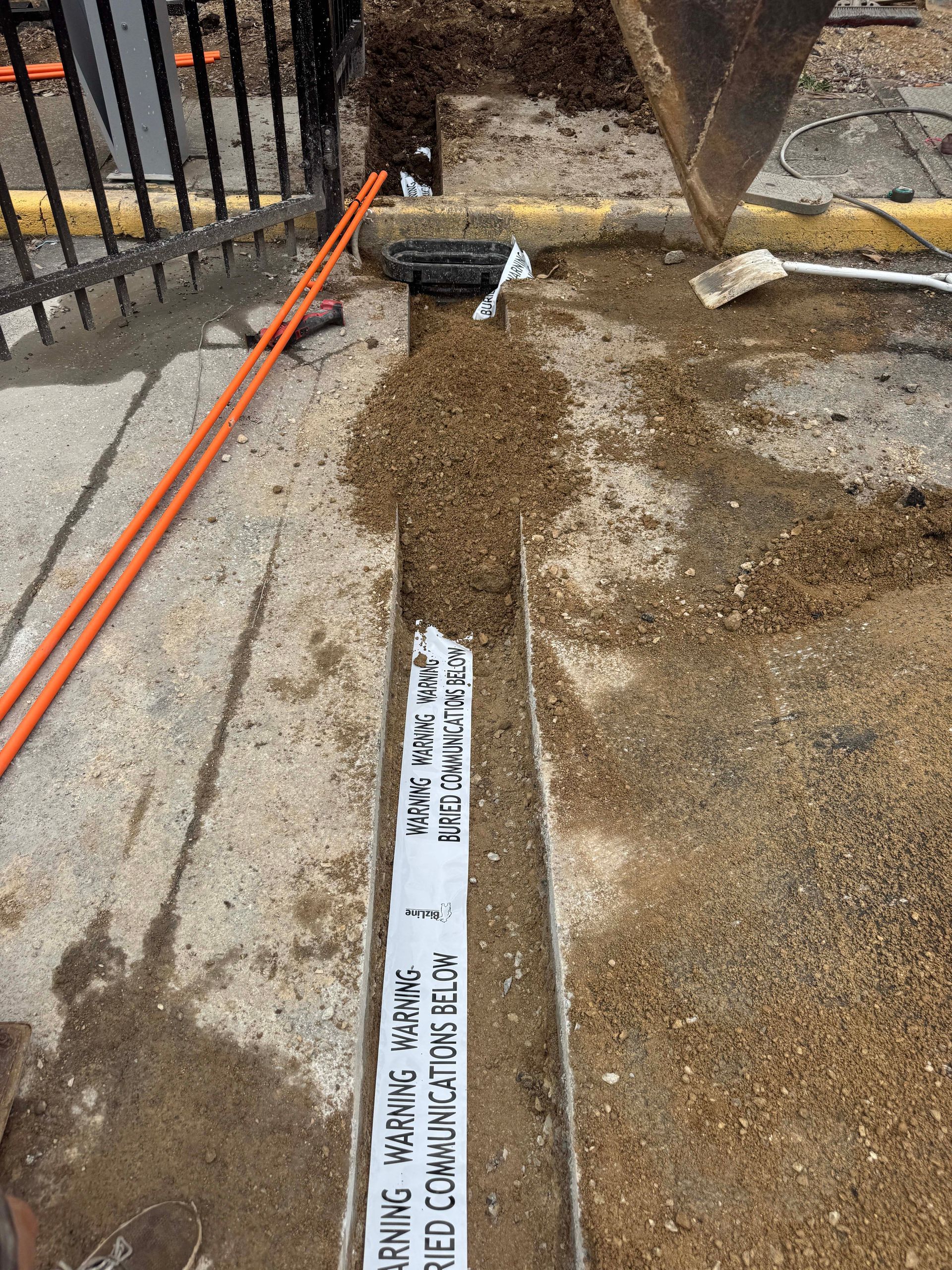 Trench dug in concrete, exposing buried utility lines. Orange conduits are visible.