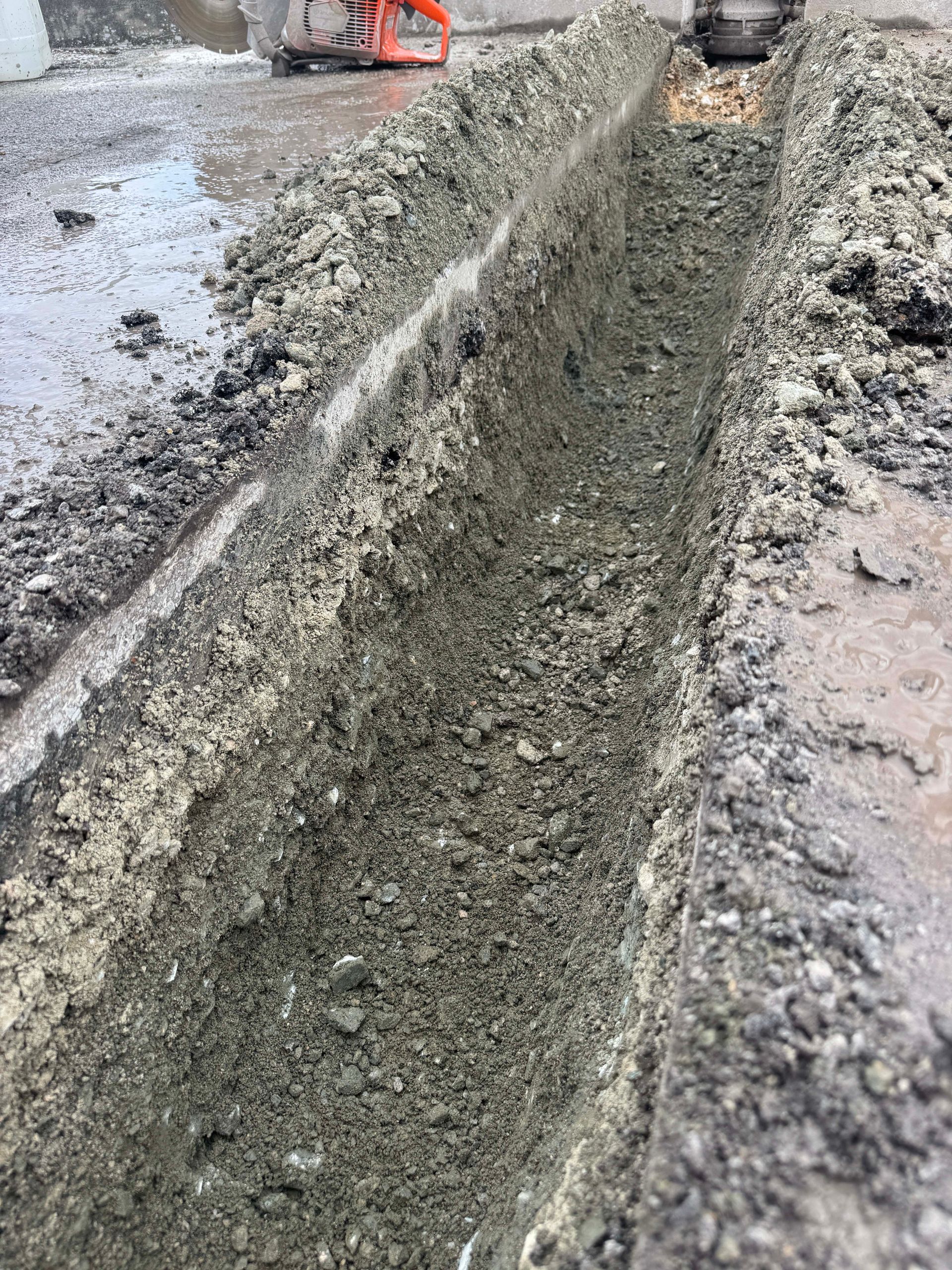 A narrow trench cut into wet asphalt, likely for utility work. Dark gray asphalt and gravel fill the trench and surround it.