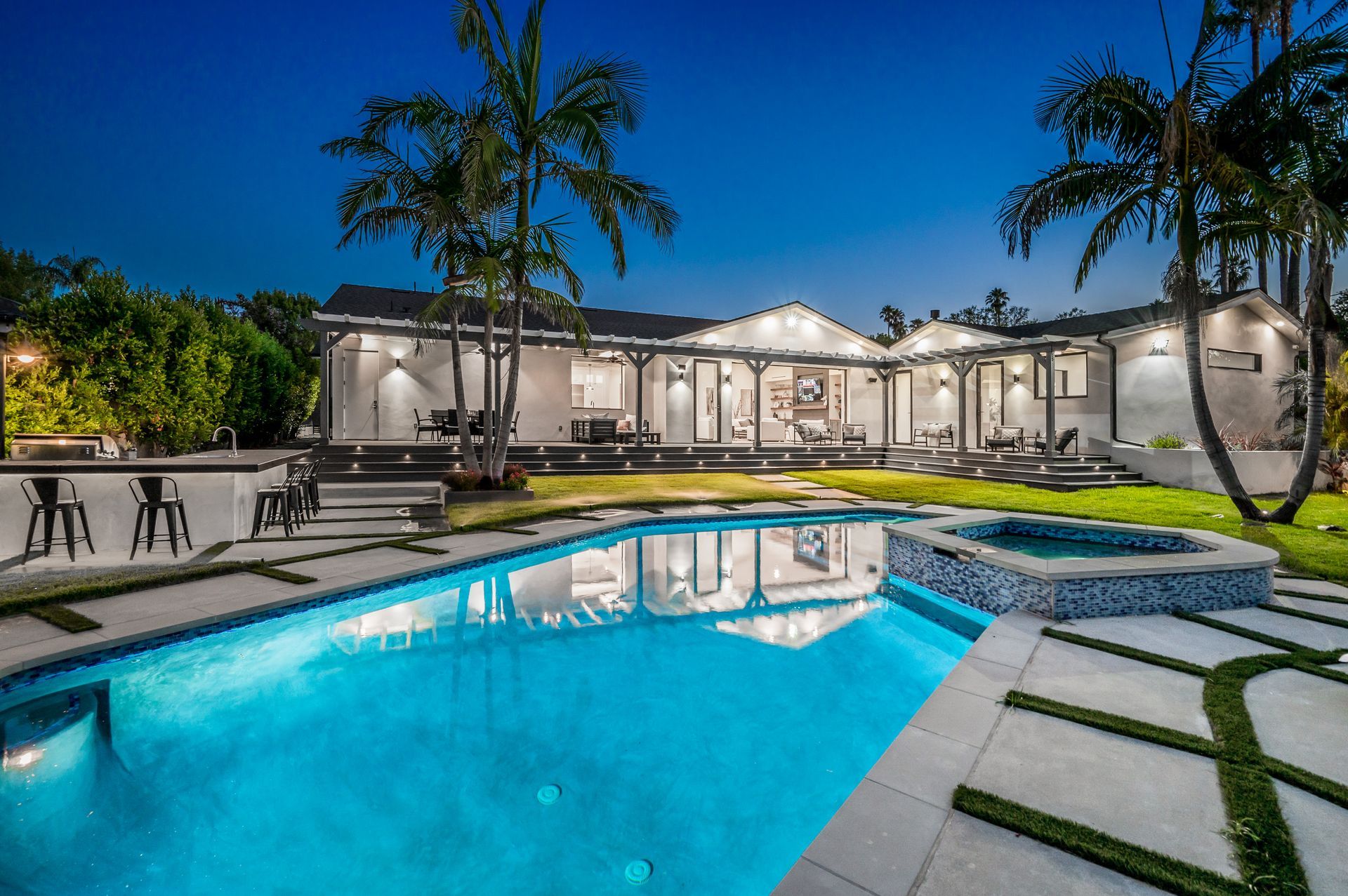 Modern house with lit backyard pool at dusk, palm trees, and a paved patio