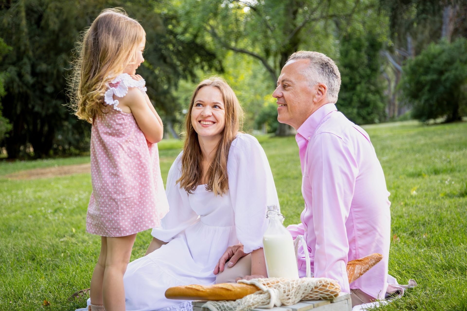Three people picnic on grass, smiling in a sunny park with flowers and bread nearby