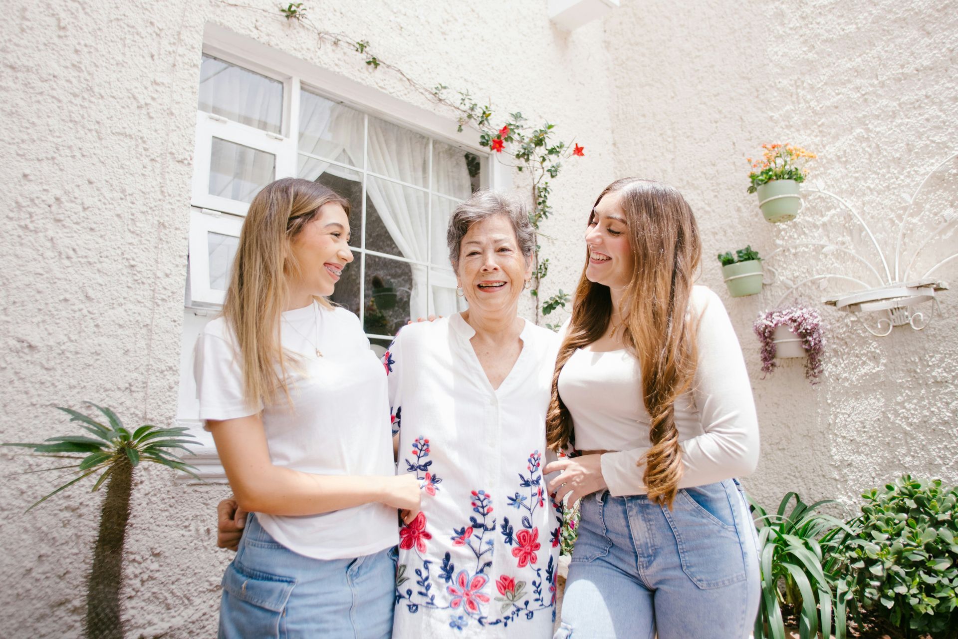 Three women smiling, hugging in front of a white building with plants.