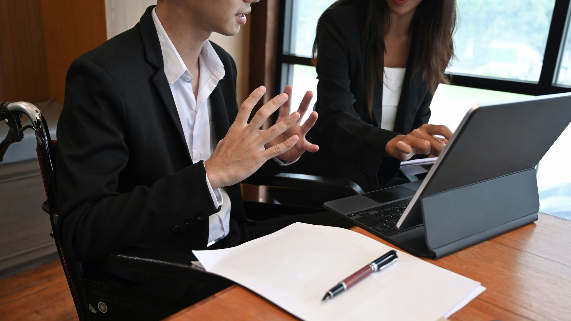 Two people in suits at a wooden table; one points a pen, the other takes notes next to a tablet.