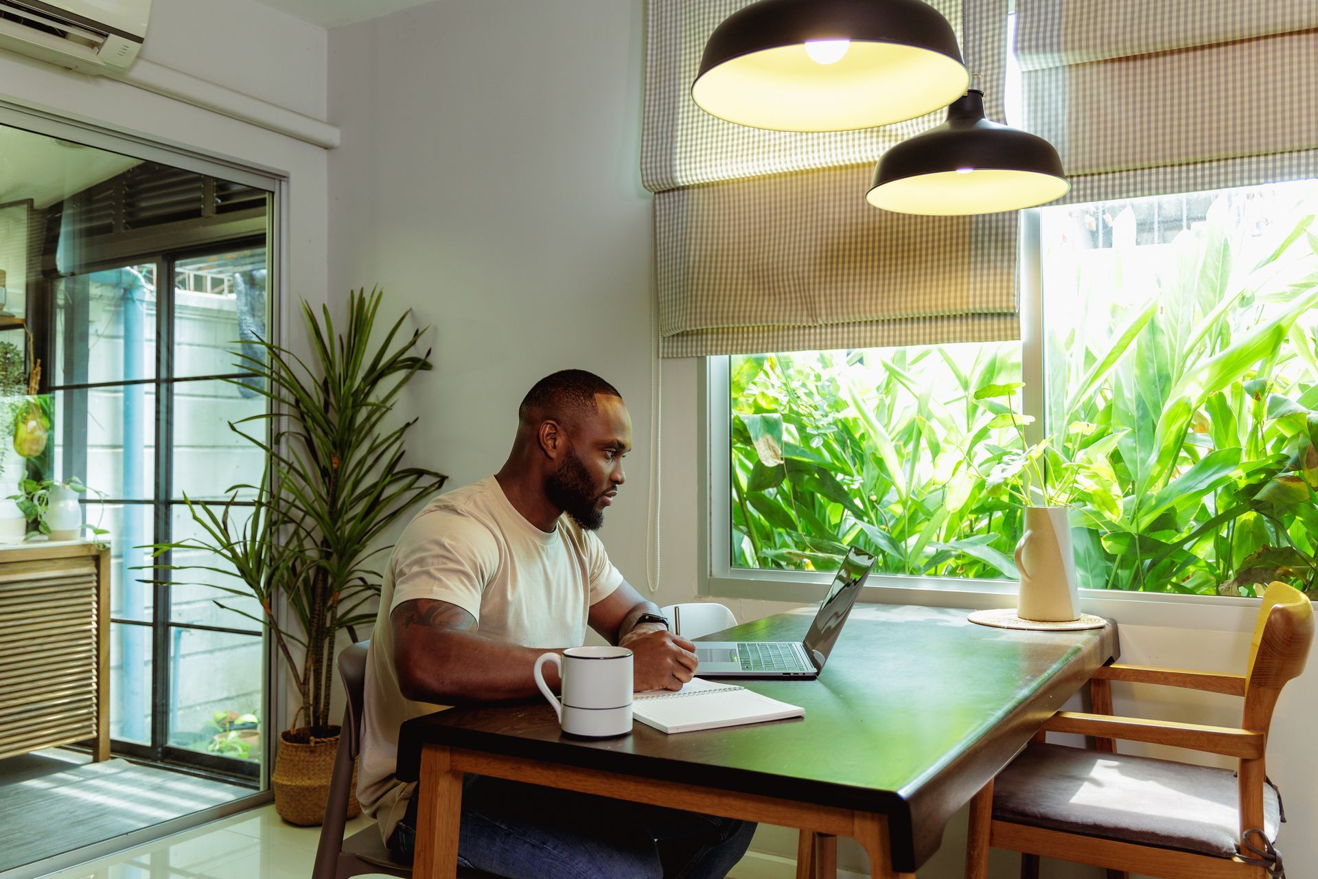 Person sitting at a dining table with a mug, working on a laptop in a bright room with large windows and plants