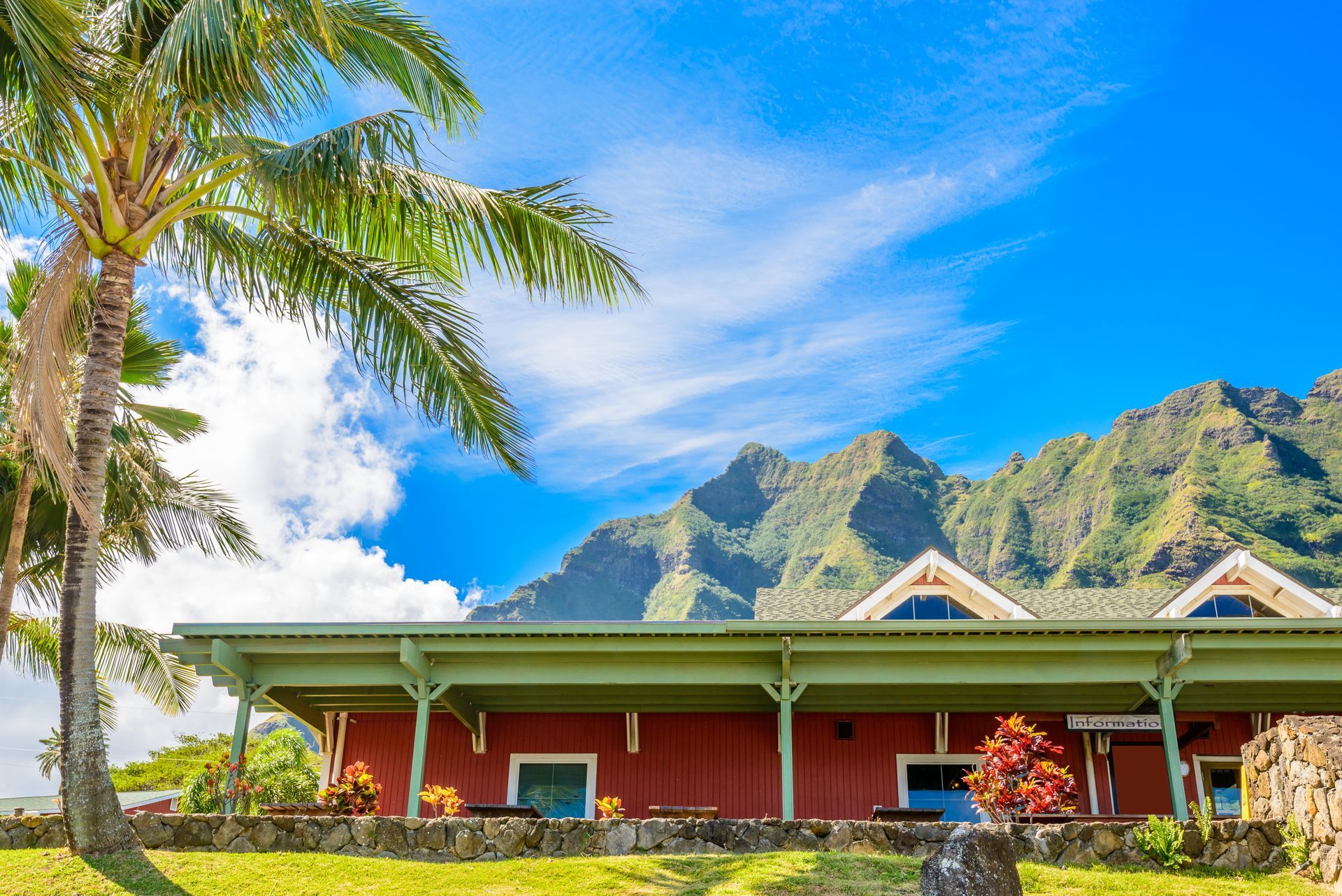 Tropical resort building with red walls, palm trees, and green mountains under a bright blue sky