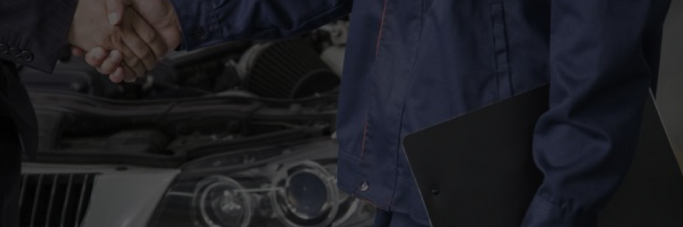 Person in a blue jumpsuit stands by a car with the hood open, holding a black folder.