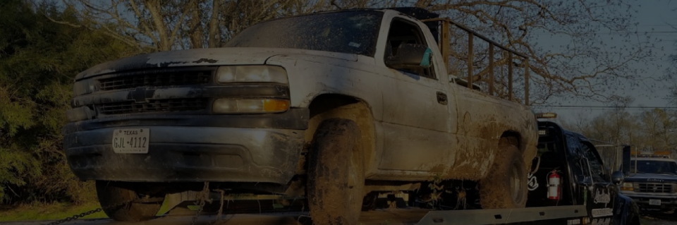 Mud-covered silver pickup truck loaded on a flatbed tow truck in a roadside setting