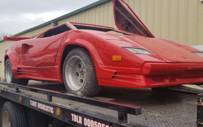 Red sports car on a car trailer, parked in front of a beige building with its hood raised.