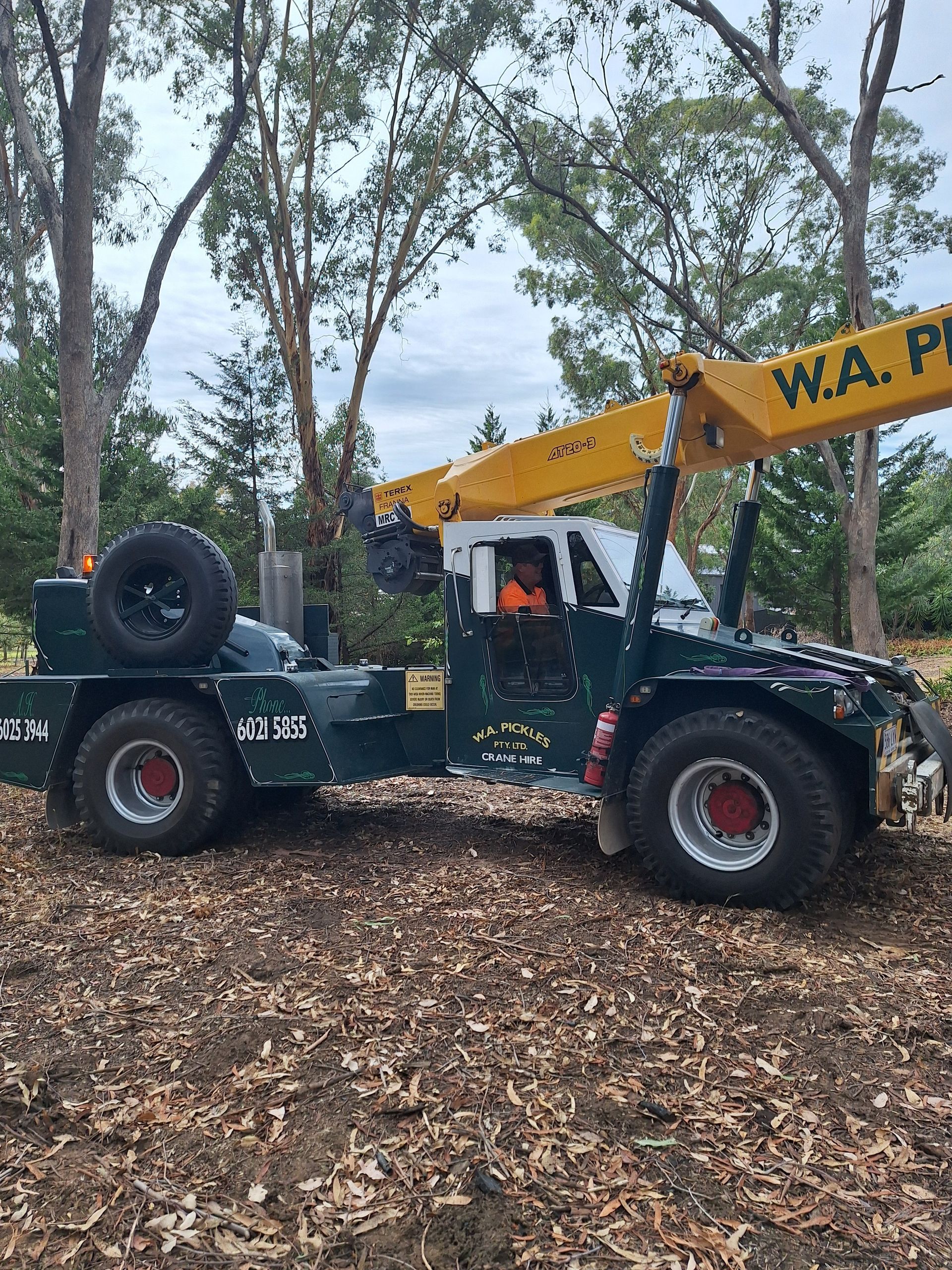 Yellow excavator on a flatbed truck in a wooded area with fallen leaves on the ground.