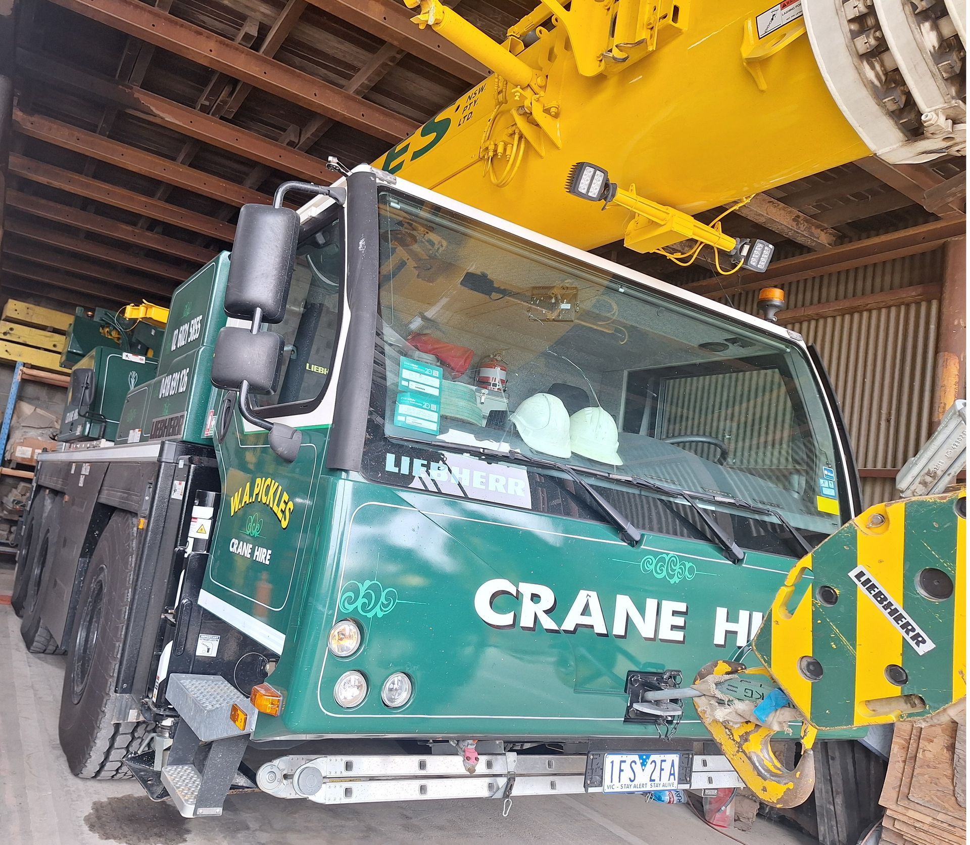 Green crane truck in a warehouse, with a yellow crane arm overhead.