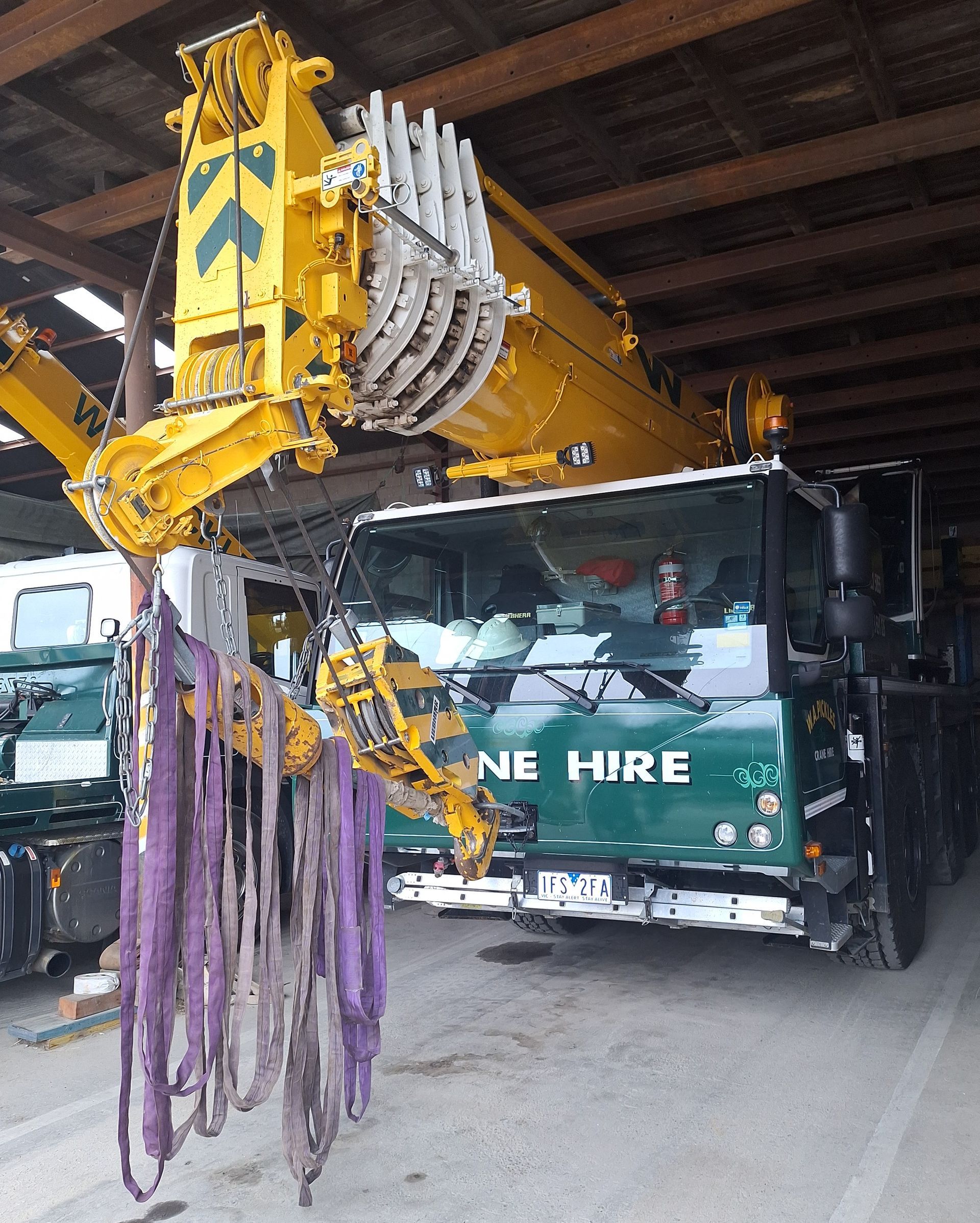 Yellow crane arm lifting a black truck with “NE HIRE” logo in a warehouse, with purple straps hanging nearby