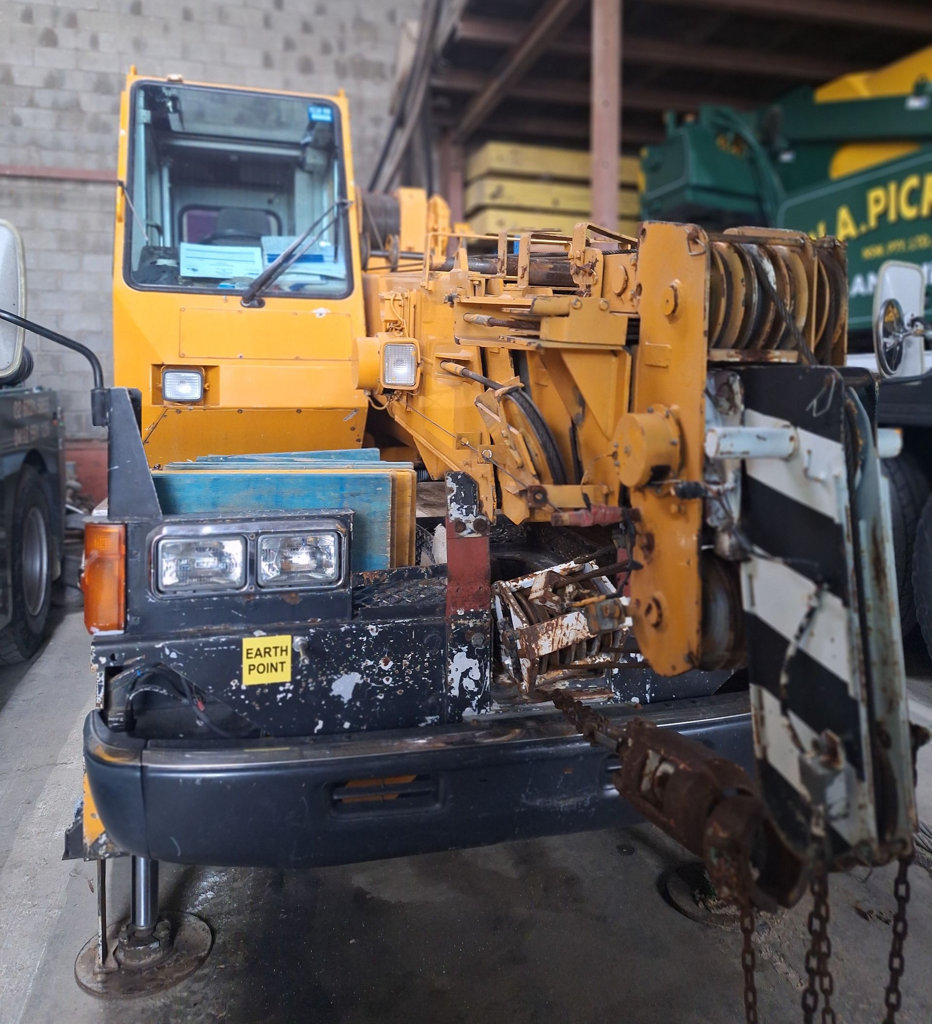 Yellow crane truck in a warehouse, with a damaged front and a large machine attached to its side.