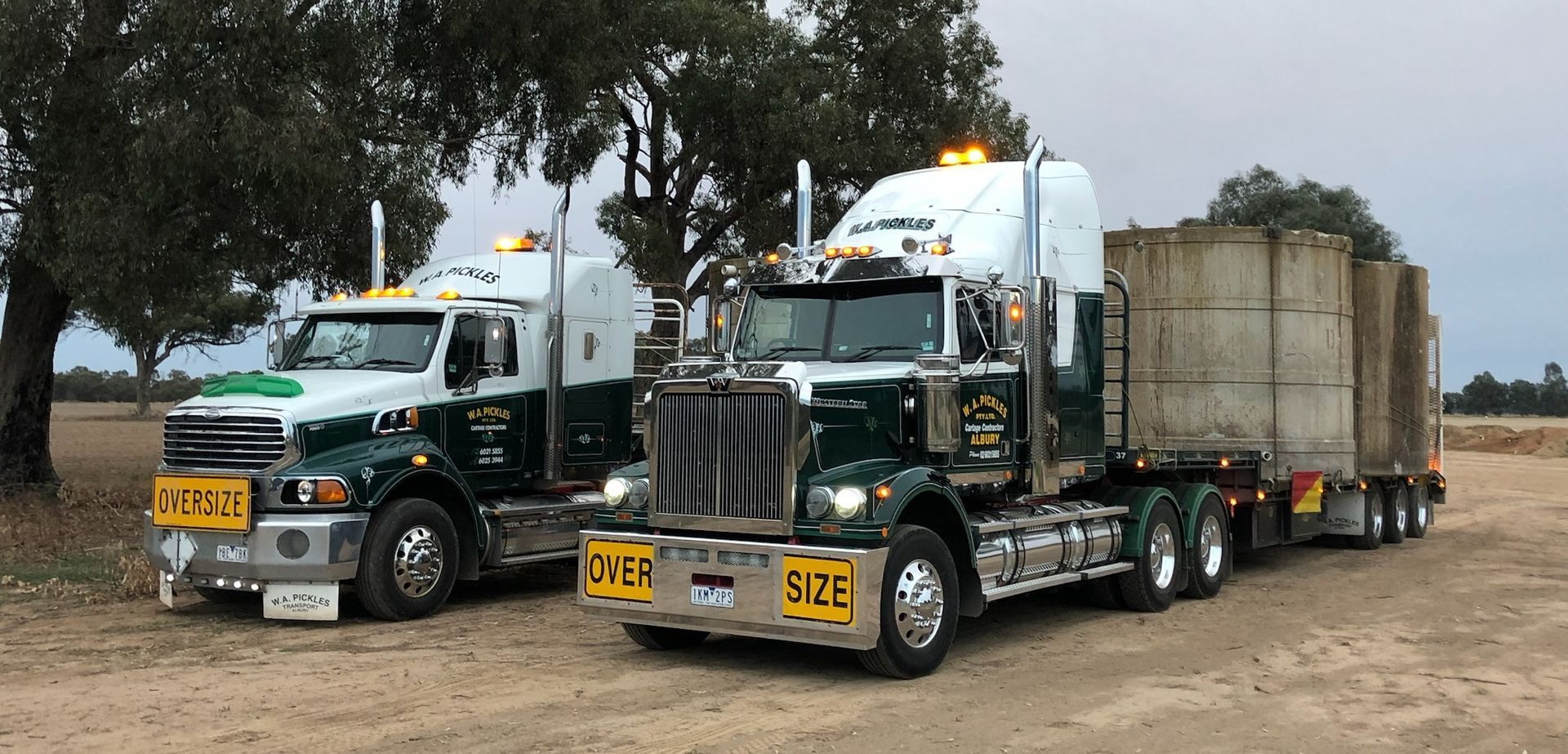 Two dark green semi trucks parked side by side on a dirt lot, each hauling large loads.