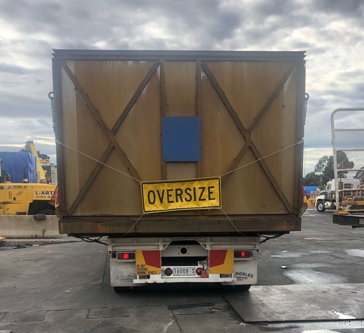 Rear view of an oversized cargo load on a trailer, with a yellow “OVERSIZE” sign and cloudy sky.
