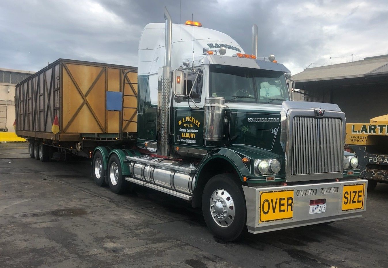 Green semi-truck hauling a covered trailer in an industrial lot, with yellow oversize load signs