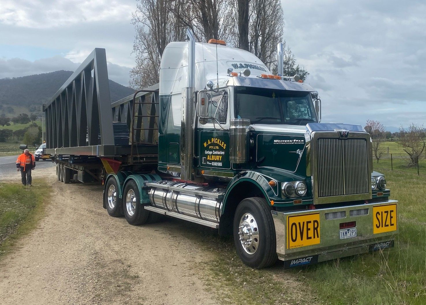 Dark green semi-truck hauling a long flatbed trailer on a rural gravel road under cloudy skies.