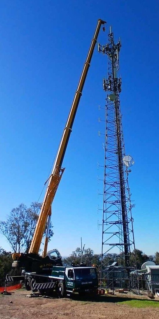 Crane lifting a tall metal tower beside parked trucks under a clear blue sky