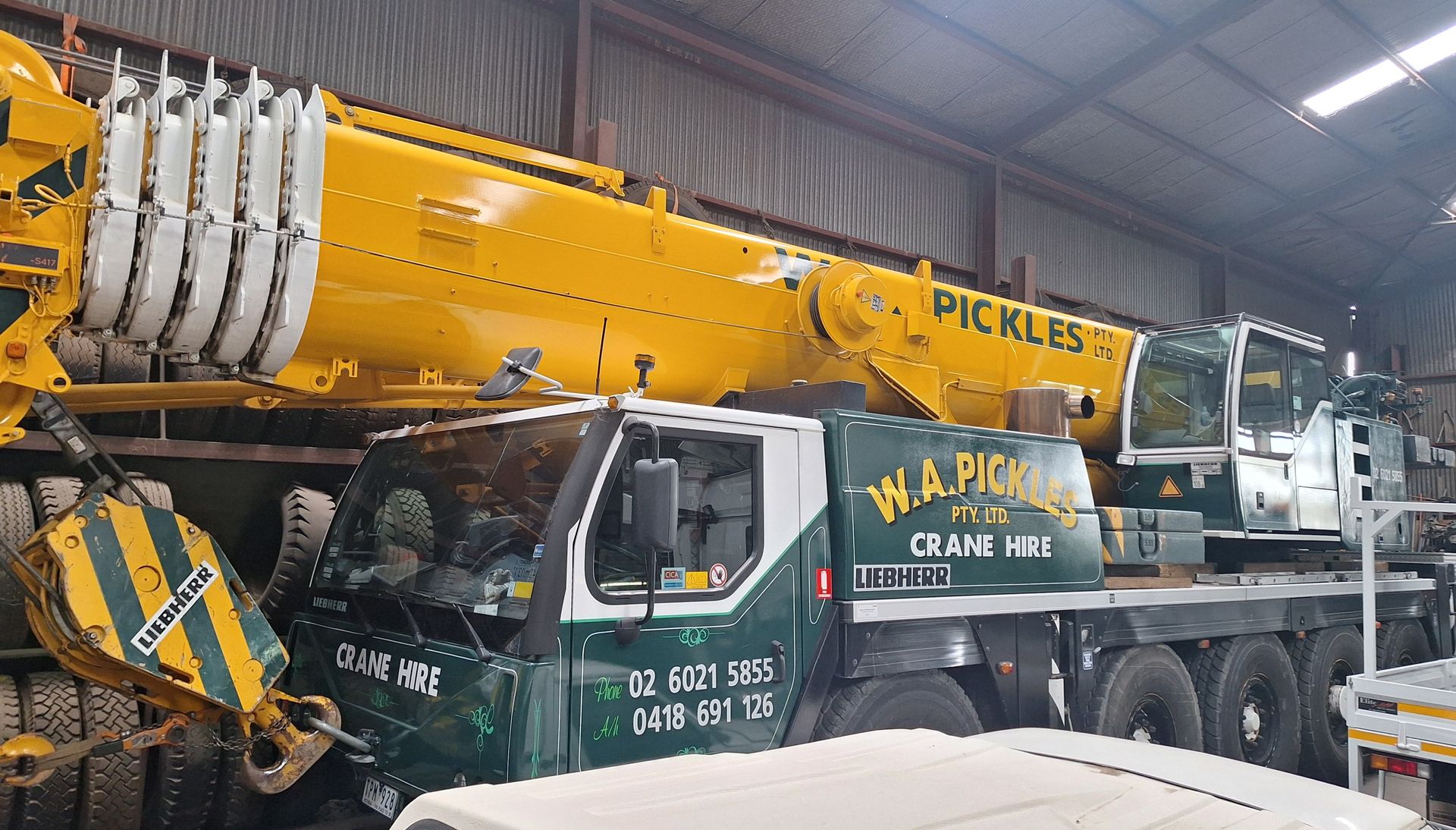 Large yellow mobile crane in a warehouse, with a green truck cab and “PICKLES” branding visible.