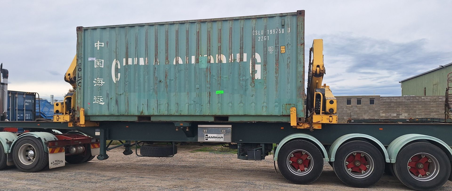 Green shipping container on a flatbed trailer with a yellow crane truck in an industrial yard