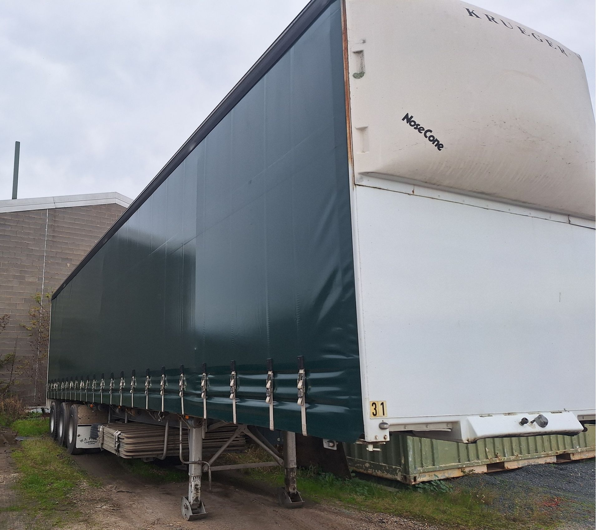 Covered semi-trailer parked outdoors on grass beside a building, with a green tarp and white front panel