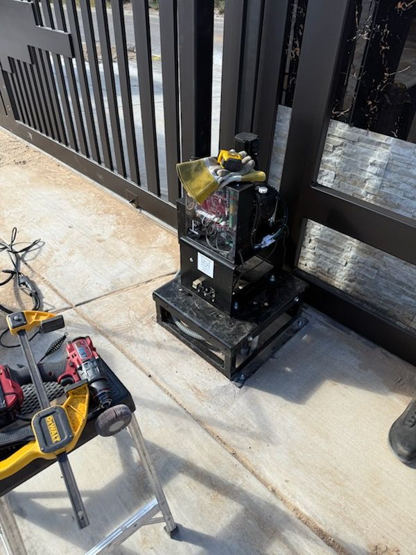 Black gate opener with yellow accents, mounted on a stand, next to a black metal gate.