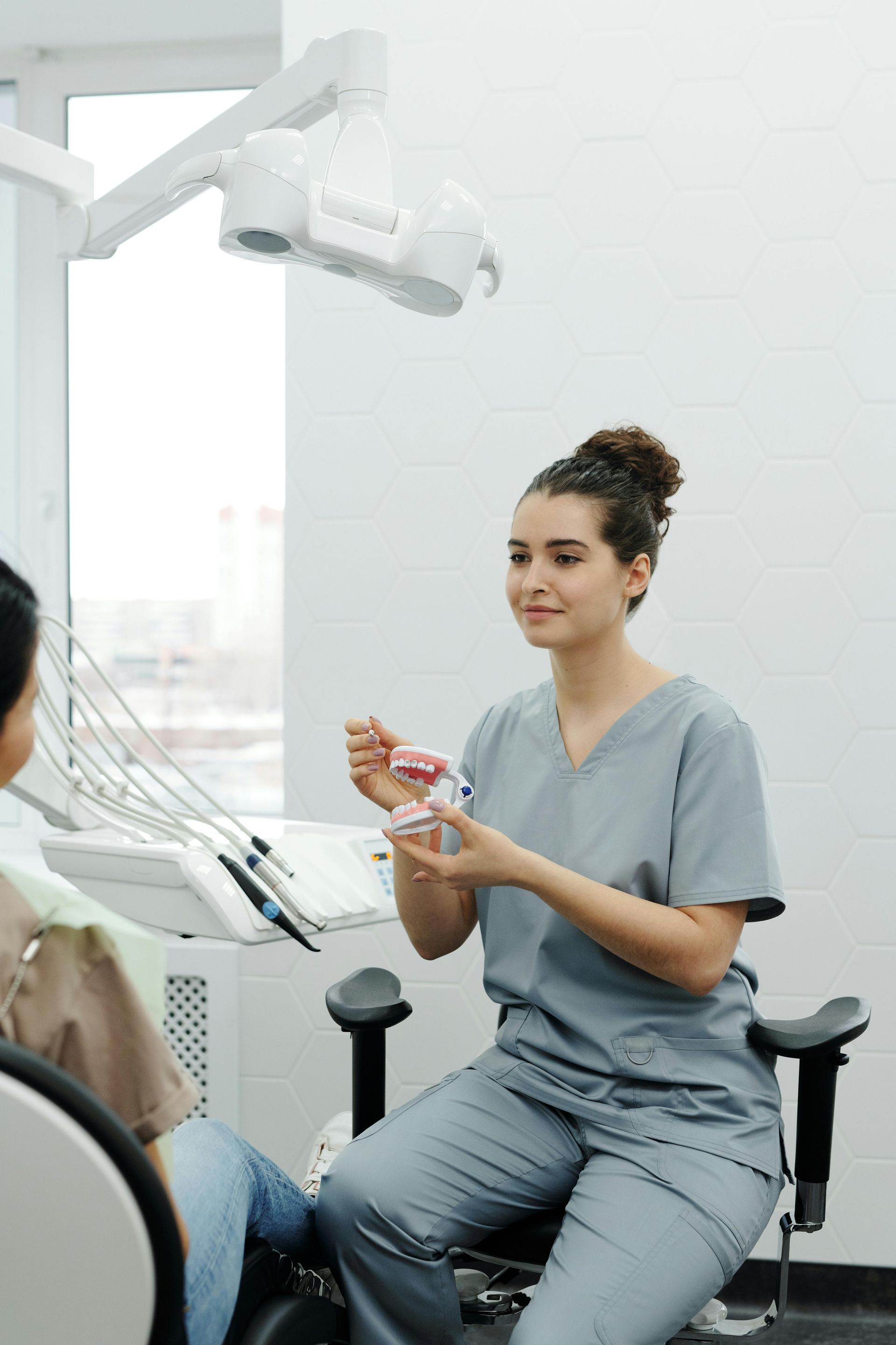 Dentist in gray scrubs holding a teeth model, explaining to a patient seated in a dental chair in a bright office.