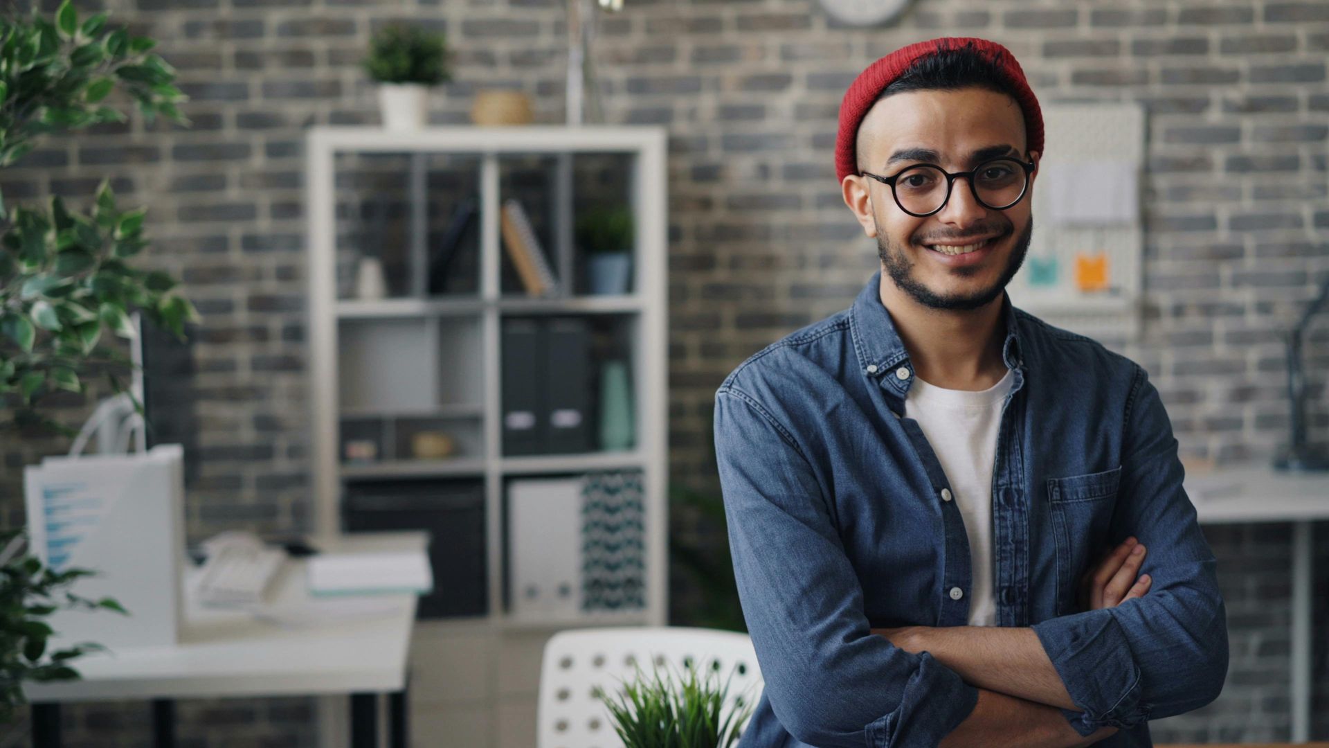 Man with glasses and red beanie smiles, arms crossed, in a modern office setting.