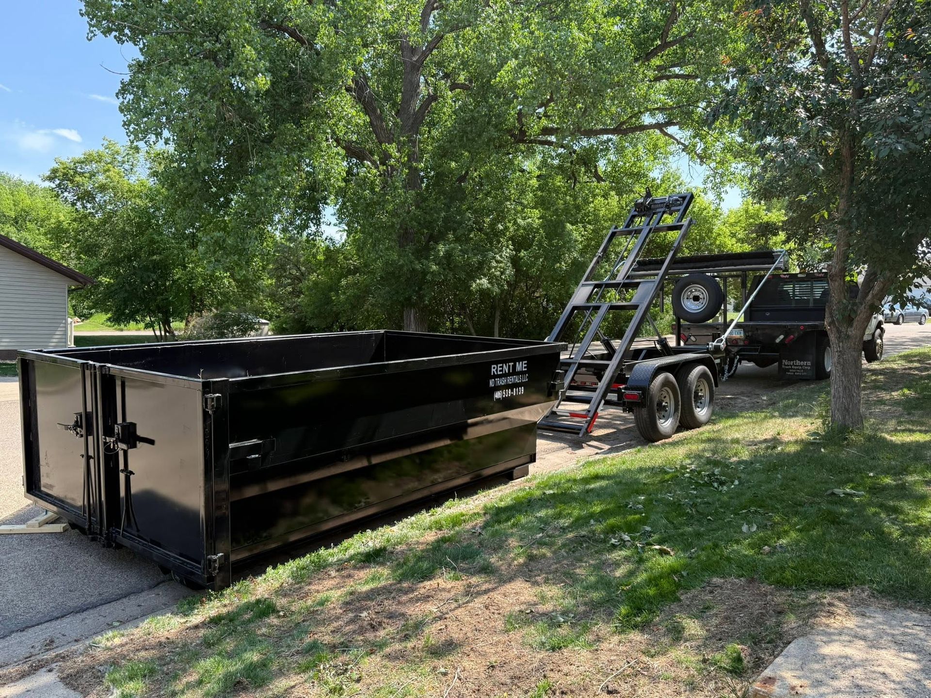 Black dumpster on a trailer beside a house, with a ladder, under trees on a grassy driveway.