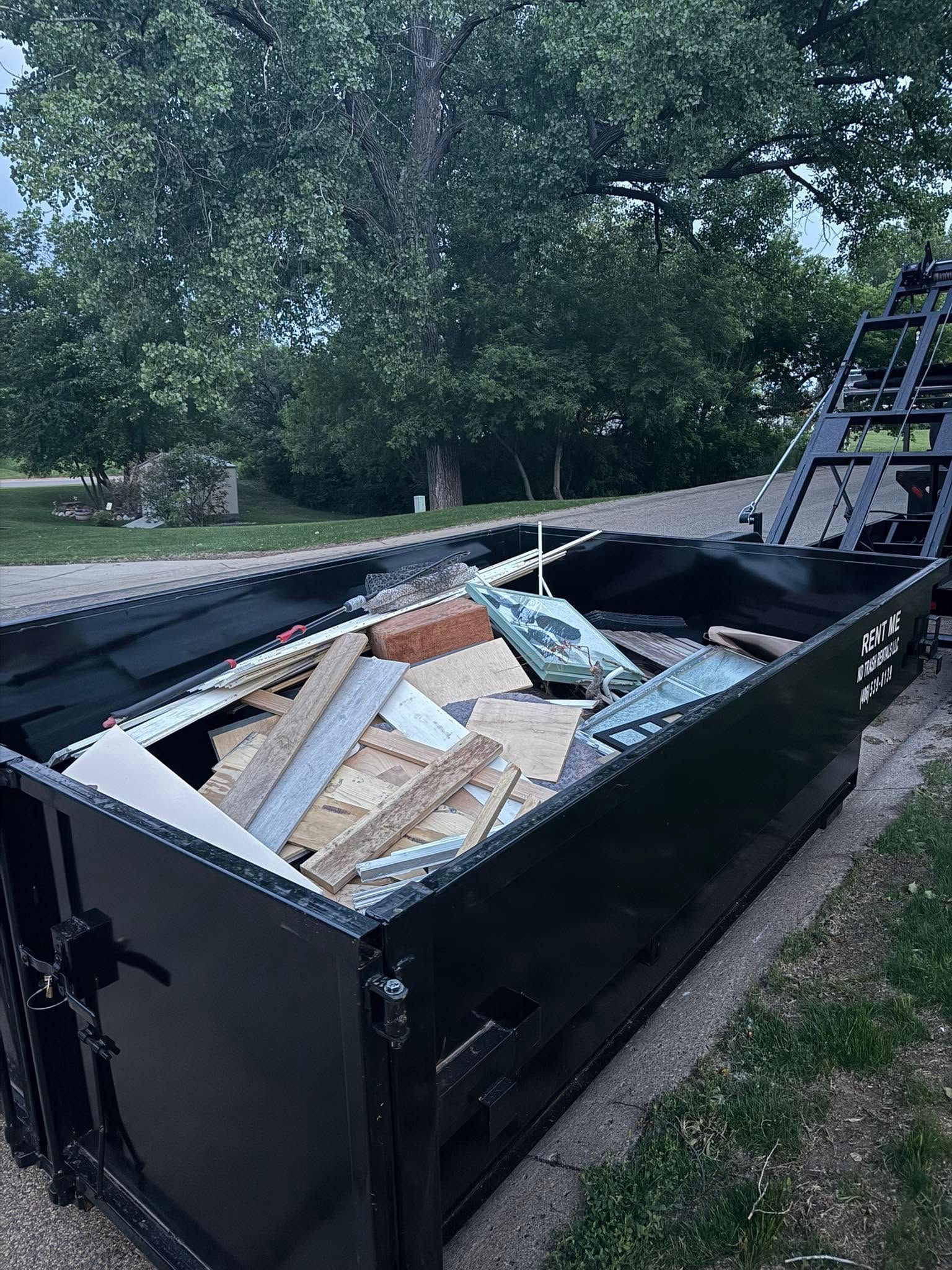 Black dumpster filled with construction debris beside a tree-lined road
