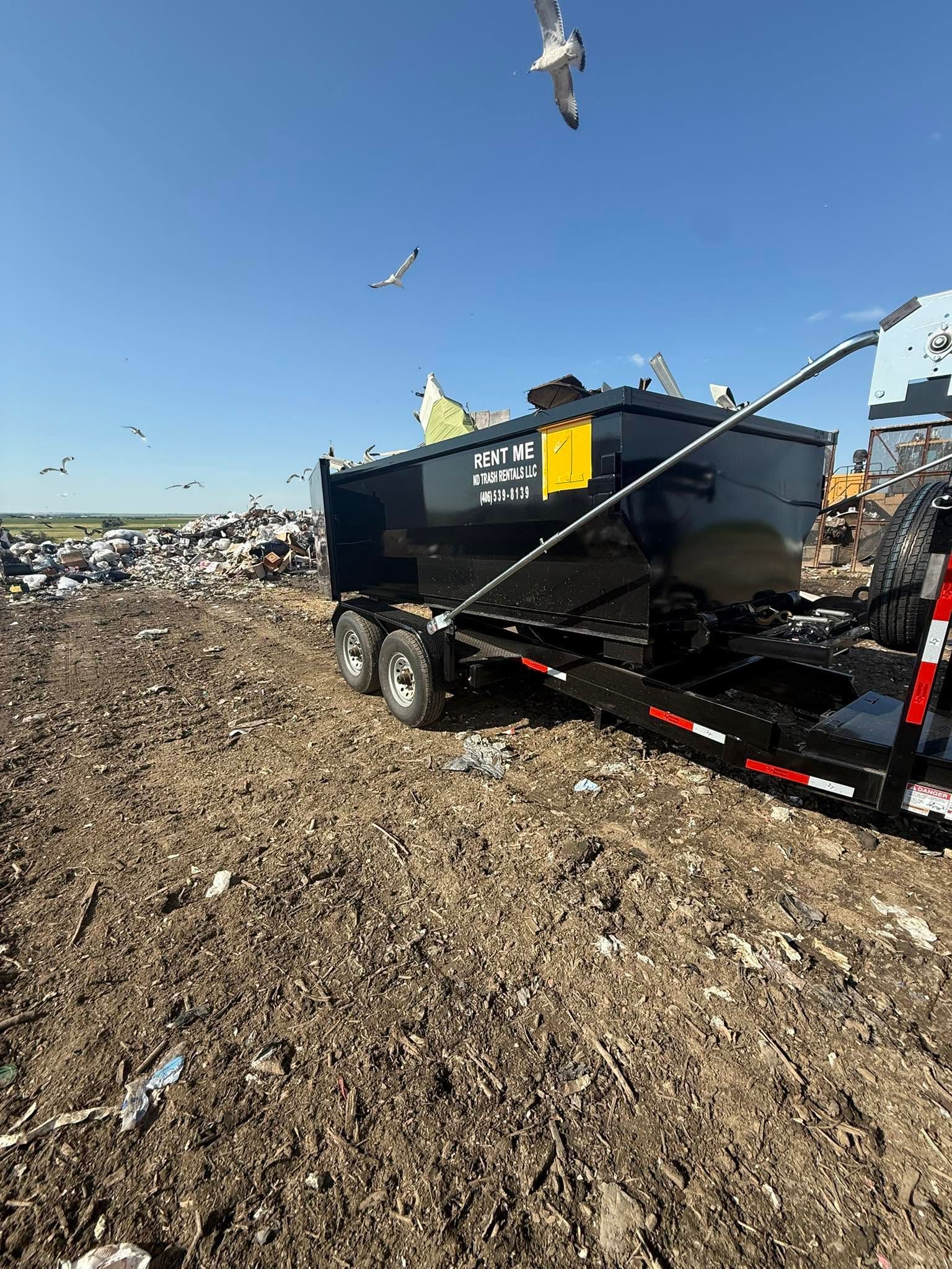 Black trailer in a muddy field under a clear blue sky, with birds flying overhead