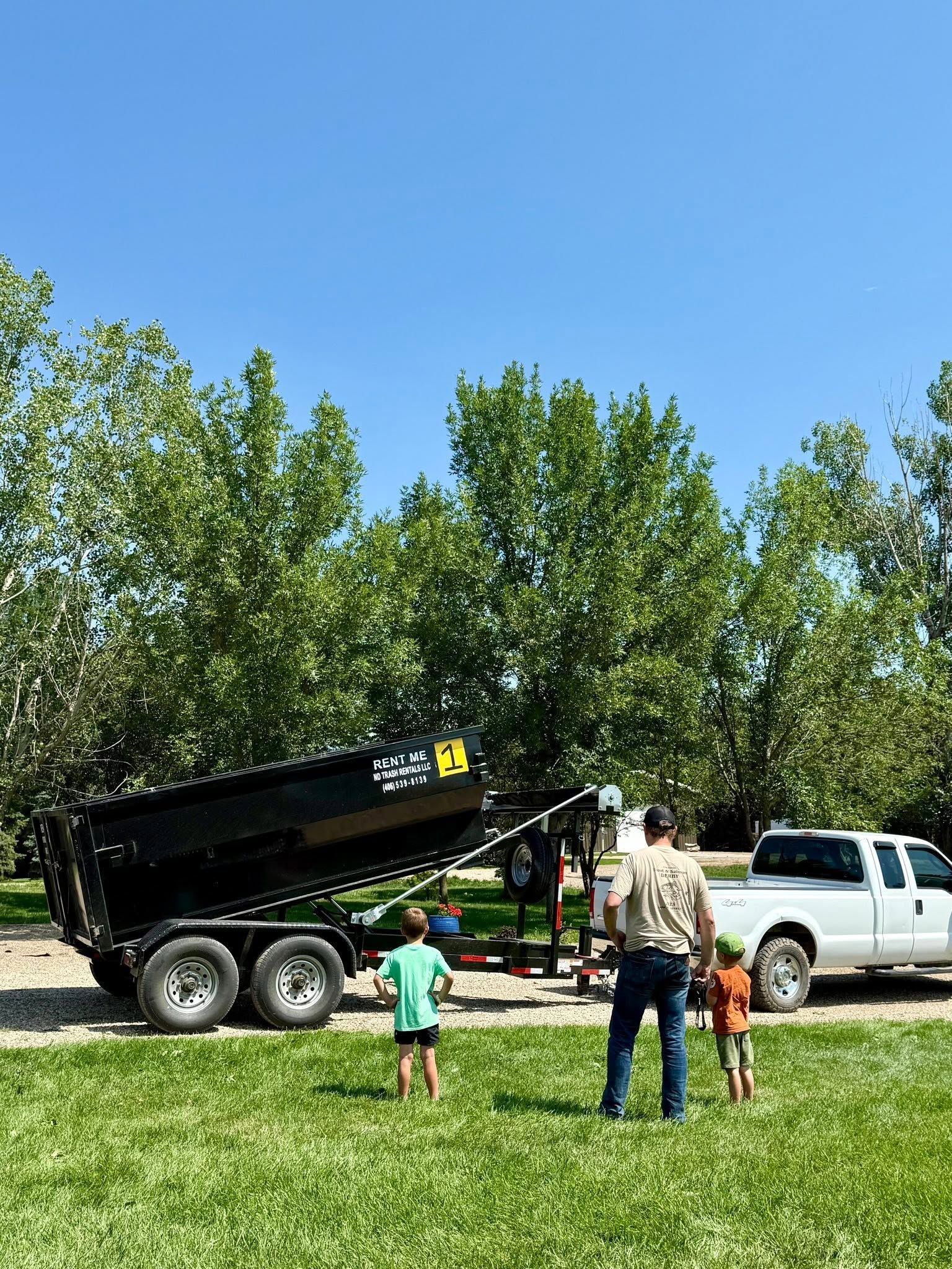 Black dump trailer with raised bed beside a white pickup; two children and an adult stand outside on a sunny day.