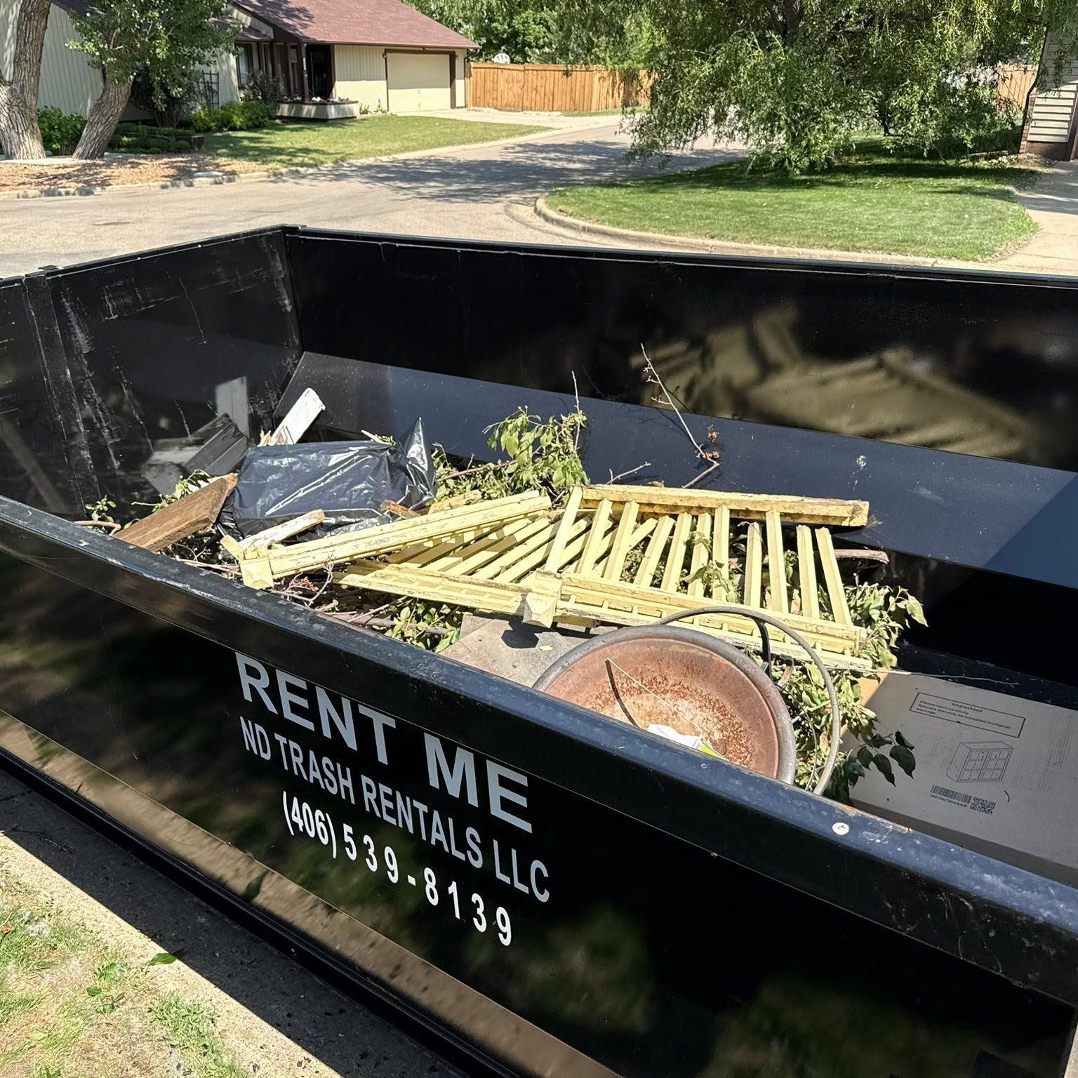 Black dumpster filled with yard debris and broken wood in a suburban driveway