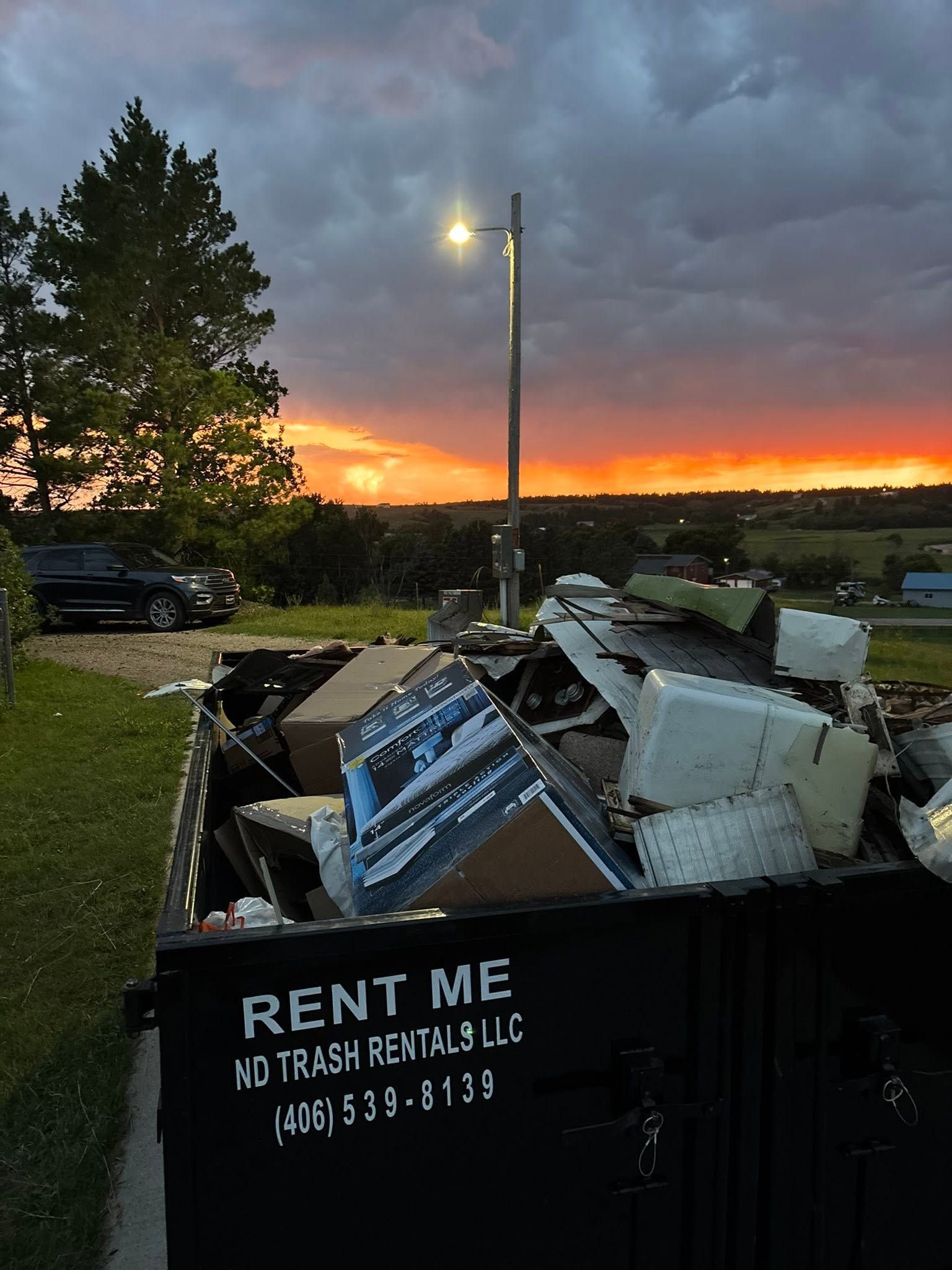 Dumpster full of scrap appliances at sunset, with a tall lamp post and trees in the background