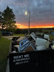 Dumpster full of debris at dusk, with a lit pole and orange sunset sky in the background
